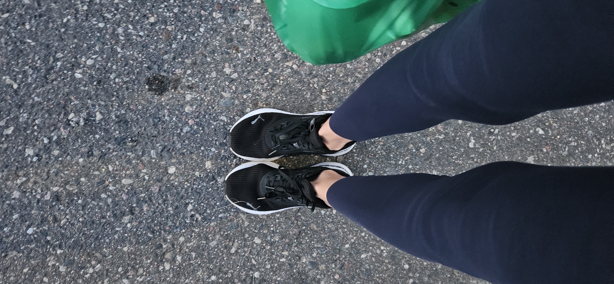 This is a top-down, eye-level shot of a person standing on a textured asphalt surface. The person is wearing black athletic shoes with white soles and white laces, and dark blue leggings. A portion of a bright green object, possibly a bag or piece of clothing, is visible on the right side of the frame. The background is the asphalt surface, which is grey and speckled with small stones and pebbles. The lighting suggests it is daytime. No specific landmarks of Vitsebsk, Belarus are visible.