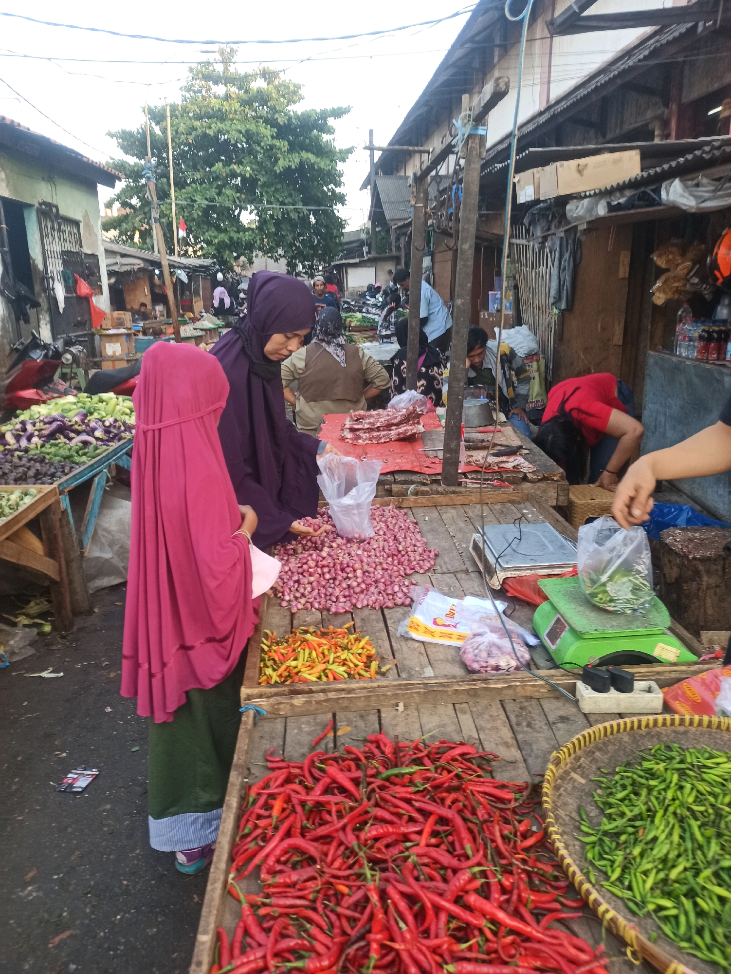 The image depicts an outdoor market scene in Cikarang, Indonesia. The foreground is dominated by wooden stalls laden with fresh produce, primarily chili peppers of various colors and sizes, and red onions. A woman in a purple hijab and a person in a pink hijab are interacting with the produce, with the woman in purple appearing to be selecting red onions from a pile. Behind them, more market stalls are visible, with various goods and other people engaged in their activities. The market is characterized by its rustic wooden structures, corrugated metal roofing, and a general atmosphere of a bustling local marketplace. Overhead, wires and cables crisscross the scene, indicating the presence of electrical infrastructure. In the background, a large tree stands prominently, adding a natural element to the urban market setting. The lighting suggests it is either early morning or late afternoon.