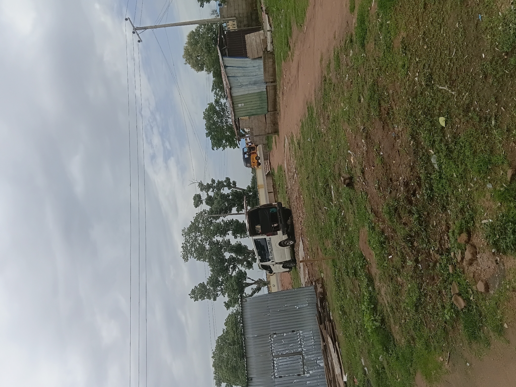 The image depicts a scene in Jalingo, Nigeria. The orientation of the image is rotated 90 degrees clockwise. In the foreground, a dirt road is visible, bordered by overgrown green grass and scattered debris. A corrugated metal structure, possibly a shed or part of a building, occupies the lower left portion of the frame. Beyond this, a white van is parked, with its rear doors open. Trees with dense green foliage are present behind the van and further into the background. A yellow auto-rickshaw is also visible further down the road.
In the middle ground, a series of structures, appearing to be makeshift buildings with corrugated metal roofing and walls, are lined up. A utility pole with multiple wires extends vertically through the scene, indicating the presence of electricity infrastructure. The sky is overcast with scattered clouds, suggesting a cloudy or potentially rainy day. The lighting is diffused, indicating it is likely daytime, though the exact time is unclear due to the cloud cover. The overall impression is of a somewhat underdeveloped or rural area with signs of informal settlement and basic infrastructure. There is some visible text on the side of the white van, which reads "THE TRAVELER'S CHOICE".