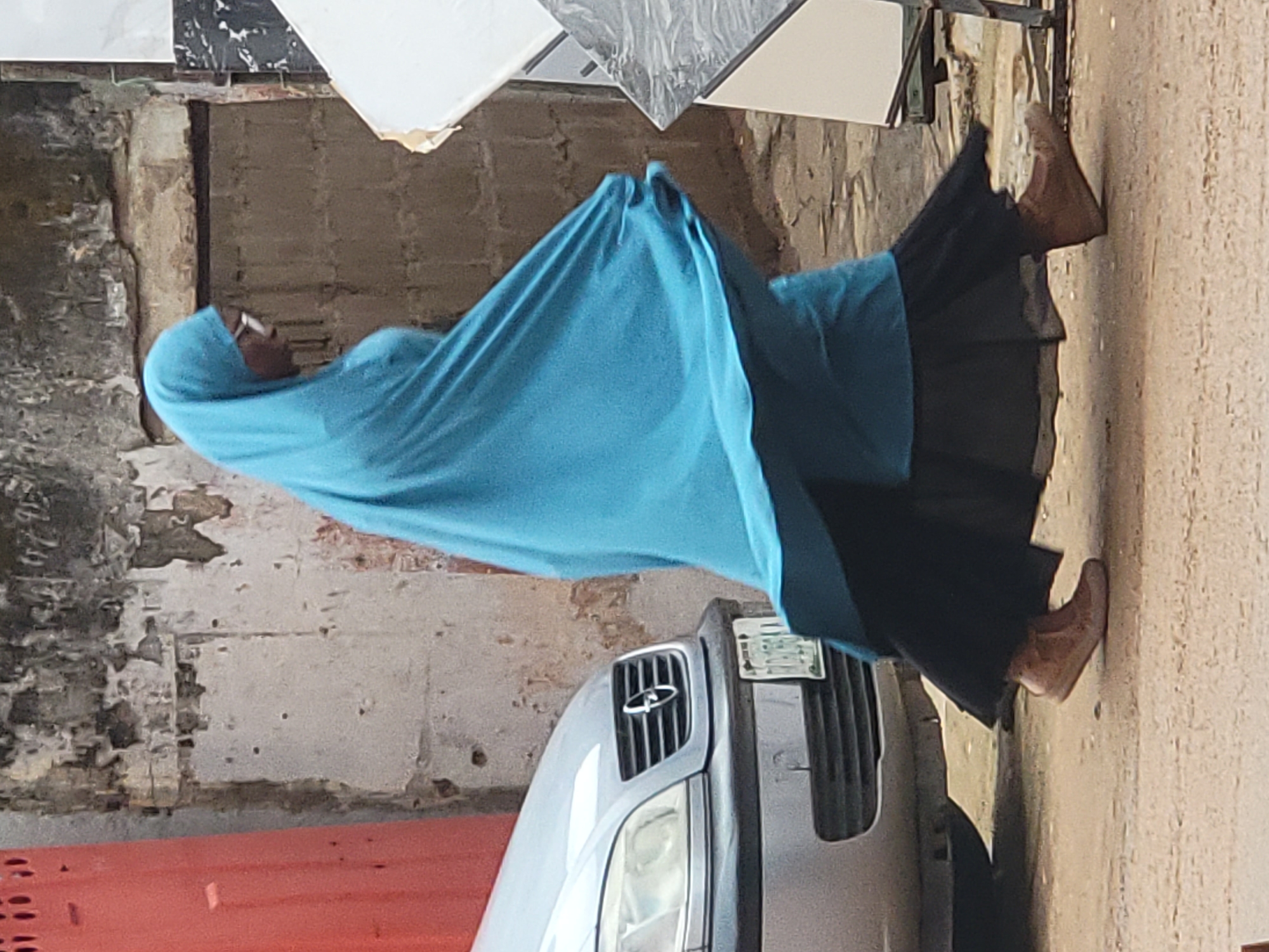 The image displays a person walking outdoors in what appears to be a street or alleyway in Maiburiji, Nigeria. The individual is dressed in a light blue head covering (hijab or similar garment) that covers their head and upper body, paired with a dark, flowing skirt or dress. They are wearing light brown shoes or boots. Only a small part of their face, including a chin and what appear to be glasses, is visible beneath the head covering.
In the foreground, partially visible on the right, is the front section of a silver or grey car, showing its grille, a headlight, and a license plate. The text on the license plate is not clearly legible. The background features textured walls, varying in shades of brown and grey, which appear aged or unfinished. Stacked against a wall in the upper part of the image are several building tiles, some with black and white patterns, and others in plain light colors, suggesting the presence of a building material vendor or a construction site.
The lighting indicates daytime, with soft and diffused illumination, possibly due to an overcast sky, as there are no harsh shadows. The setting, with its functional architecture and daily activity, portrays a common scene in an urban or semi-urban environment. The image captures a moment of an individual going about their day. No specific emotions can be discerned from the person.