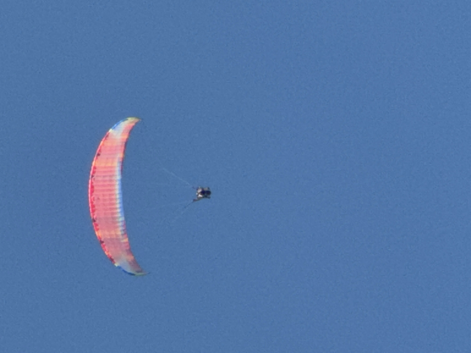 A paraglider is airborne against a clear blue sky. The paraglider's canopy is colorful, featuring stripes of pink, red, orange, yellow, and blue. A person is suspended beneath the canopy, appearing small in relation to the size of the paraglider. The image does not provide any specific details or context that would place the scene in Boreti, Montenegro, or any other specific city. The environment is solely the sky.