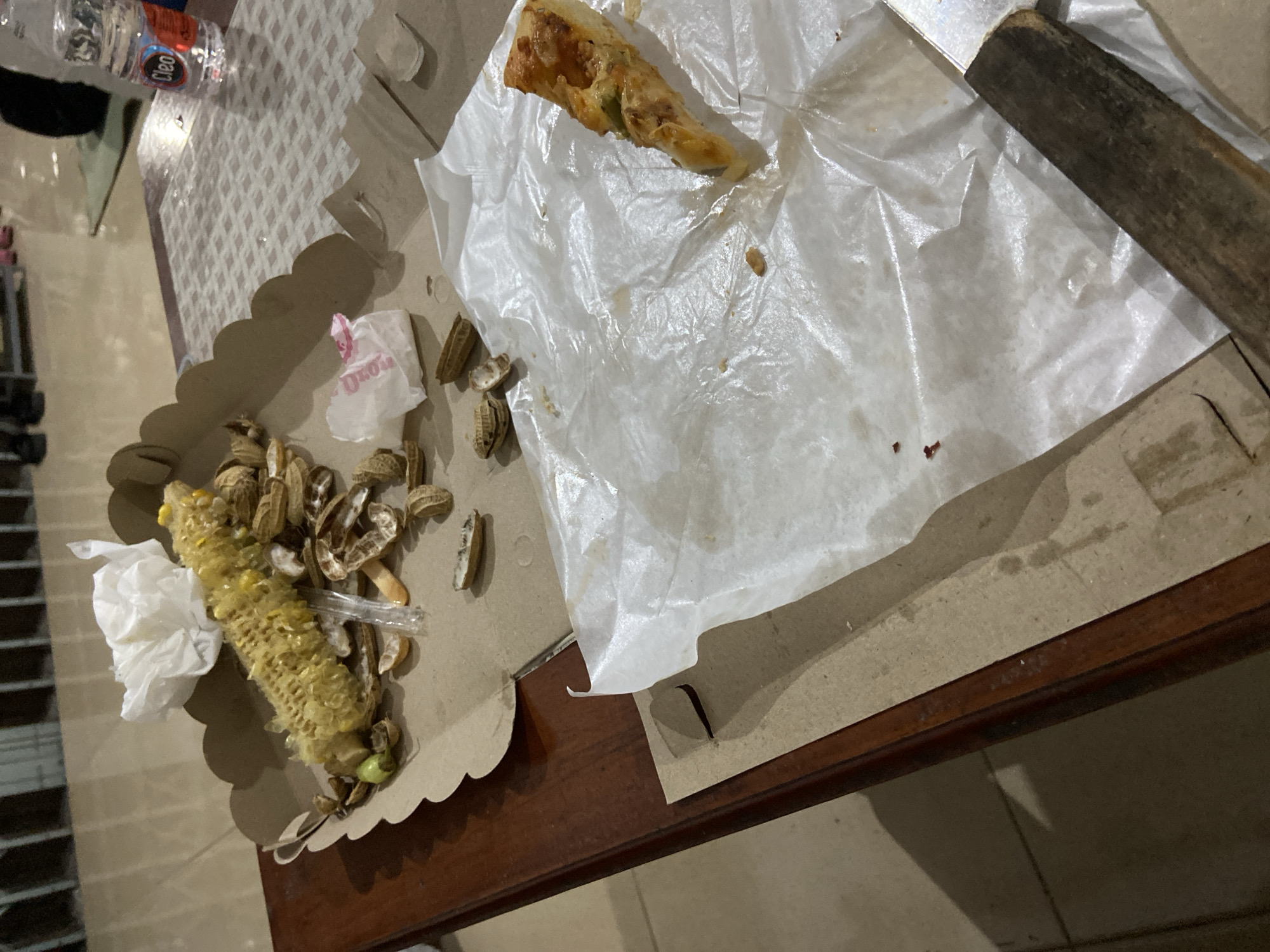 The image depicts a close-up of a table surface at what appears to be a casual gathering. On a cardboard tray, remnants of food are visible: a partially eaten cob of corn, numerous peanut shells, and a single slice of pizza resting on a sheet of white paper. A bottle of "Cleo" brand water is also present in the background. The setting suggests a post-meal environment, with discarded food and packaging indicating that consumption has concluded. The floor tiles and the wooden edge of the table provide environmental context. There is no discernible indication of the specific location within Tanjungsari, Indonesia, beyond it being an indoor space. The image focuses on the aftermath of a meal.