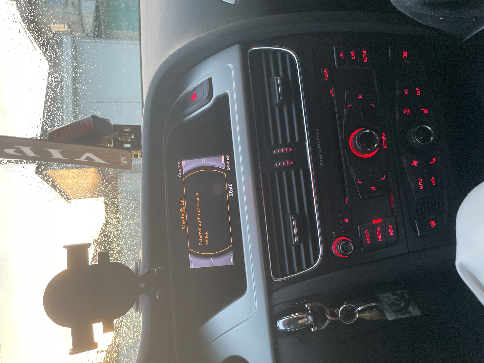 The image displays the interior of a car, specifically the dashboard and center console, with a view through a wet windshield. The primary focus is the car's infotainment system and control panel.
The central infotainment screen shows the time "20:46" and the message "External audio source is active," alongside labels such as "Media," "TP," "AUX," "Source," and "Sound." Below and to the right, the car's control panel, branded "Audi symphony," is illuminated with red backlighting. Numerous buttons and rotary knobs are visible, labeled with functions like "RADIO," "MEDIA," "INFO," "LOAD," "TEL," "CAR," "SETUP," "AC," "OFF," "RETURN," and "AUTO." Air vents are also integrated into the dashboard.
On the left side of the frame, a phone mount is attached to the windshield. A small red object inscribed with "HOLY BIBLE" rests on the dashboard, next to a "VIP" sign which partially reveals the word "CARIBI." A key fob with keys is visible in a compartment below the control panel.
The windshield is covered in numerous water droplets, indicating rain or recent precipitation. The exterior background, viewed through the wet glass, is bright but indistinct, suggesting an outdoor setting during the evening hours, consistent with the displayed time of 20:46. The car appears to be stationary. No people are visible.