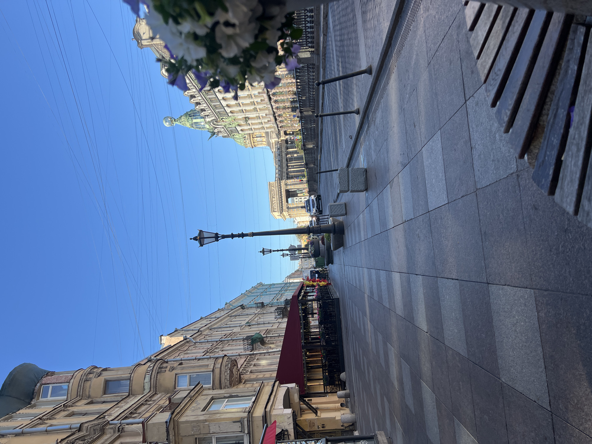 The image depicts a wide, paved street in Saint Petersburg, Russia, on a clear, sunny day. The architecture suggests a historical district, with ornate buildings lining both sides of the street. On the left, a building with a beige facade features several bay windows and decorative molding. Adjacent to it is a structure with a red awning extending over an outdoor seating area for a restaurant. Several tables and chairs are visible beneath the awning. Further down the street, more buildings with varying pastel colors can be seen.
On the right side of the street, a grand, classical-style building dominates the view. It has a large colonnade and a distinctive green dome topped with a globe-like structure. In the foreground on the right, a bench made of wooden slats is positioned on the sidewalk, partially obscuring a vase filled with white and purple flowers. The street itself is paved with large, rectangular grey stones. Lampposts, in a traditional style, are spaced along the sidewalk. A few cars are visible on the street, suggesting moderate traffic. The sky is a vibrant, cloudless blue, with a few thin electrical wires crisscrossing overhead. The overall scene conveys a sense of calm and order in a picturesque urban setting.