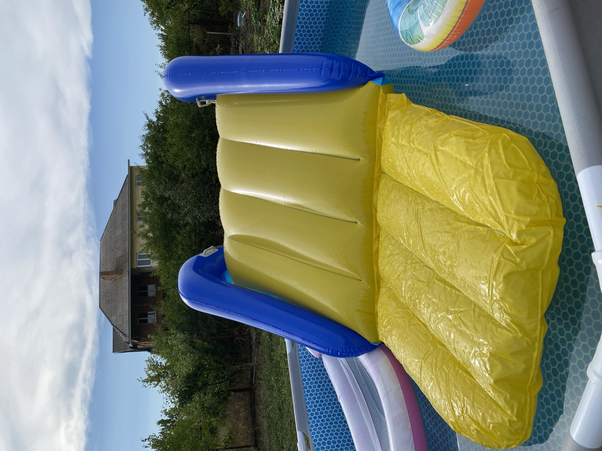 This media file captures a close-up view of a yellow inflatable slide with blue accents resting inside a blue, hexagon-patterned swimming pool. The slide appears wet, with water droplets visible on its surface, suggesting it is in use or has recently been used. The pool itself contains a colorful inflatable ring, partially submerged, and other inflatable elements are visible at the bottom of the pool.
In the background, a house with a grey tiled roof and several windows can be seen nestled amongst lush green trees. The sky above is a bright blue with scattered white clouds, indicating fair weather. The image is taken from a high angle, looking down into the pool.
The scene evokes a sense of summer fun and outdoor recreation. The location is Kropyvnytskyi, Ukraine, and the time of day appears to be daytime, given the bright sunlight. There is no visible text in the media file.