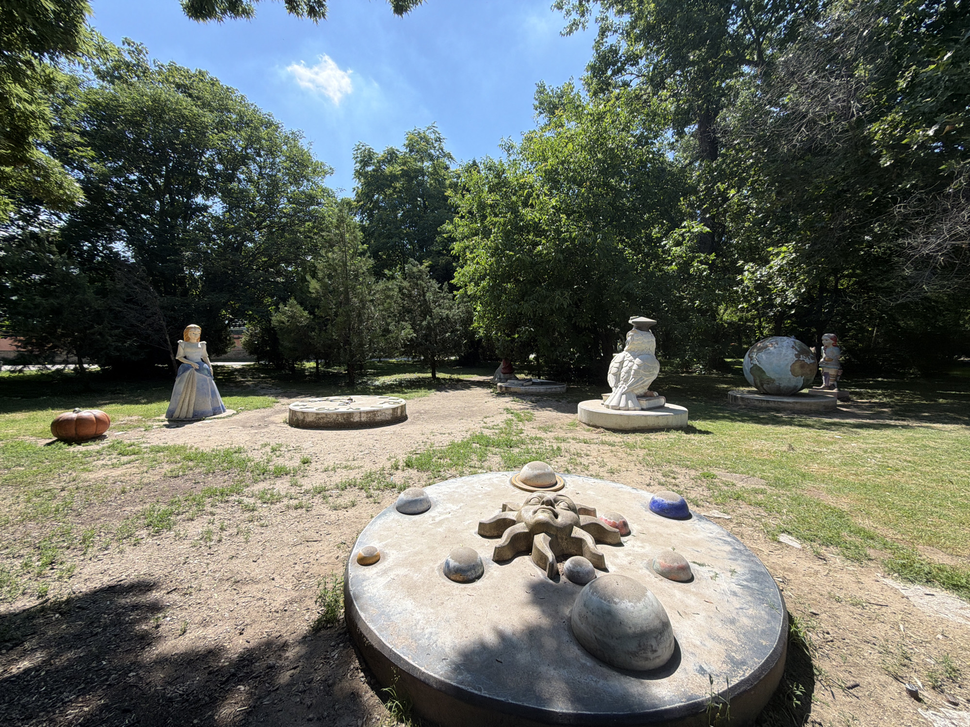 This is an outdoor daytime photograph of a sculpture park in Pazardzhik, Bulgaria. The foreground features a large, circular concrete sculpture with a depiction of a sun with a face in the center, surrounded by several spherical objects, resembling planets. In the middle ground, a statue of a woman in a blue and white dress stands near a round concrete base with a celestial map on it. To the right of the woman, a small globe sculpture and a figure of a person in a red hat are visible. Further back, a large white owl sculpture is present. The ground is covered with dirt and patches of grass. The background is dominated by a dense canopy of green trees under a clear blue sky with a few white clouds. A bright orange pumpkin sits to the left of the woman statue.