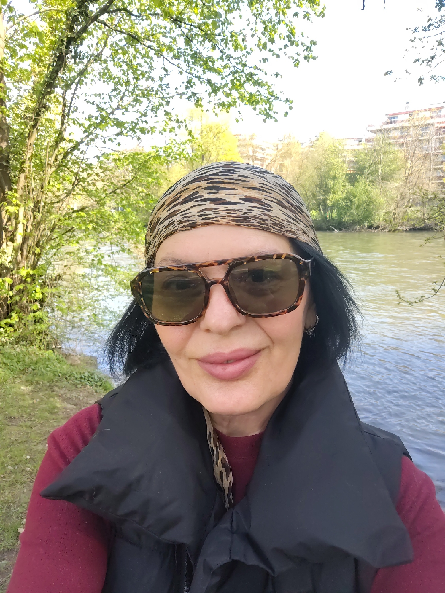 A woman stands near the Ill River in Strasbourg, France. She wears a leopard-print headscarf and sunglasses and a black life vest over a maroon sweater. The river flows behind her, flanked by trees and buildings. Sunlight illuminates the scene, highlighting the river's surface and the foliage.