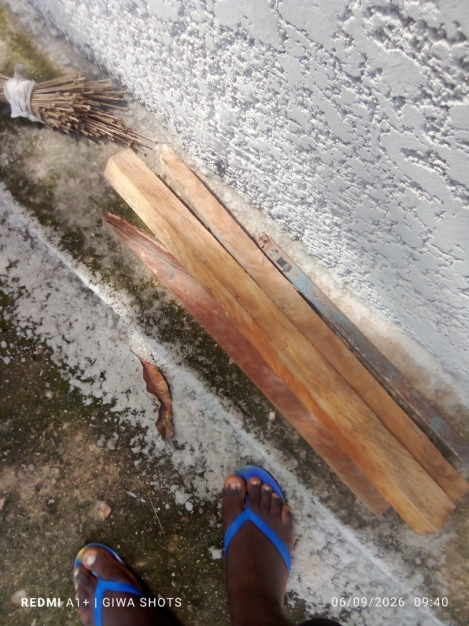 A high-angle, medium shot captures a scene with a rustic and utilitarian aesthetic. In the upper left corner, a bundle of dry, brown twigs, likely part of a broom, is tied with a white cloth. Adjacent to this, three wooden planks lie horizontally, their surfaces showing natural wood grain and some signs of wear. A slender piece of weathered blue metal or wood runs parallel to the planks. The ground beneath is a rough, grey concrete surface, stained with patches of green moss and dirt. A single dry, brown leaf rests on the concrete. In the foreground, a pair of dark brown feet wearing bright blue flip-flops are visible. The left foot is closer to the camera, with toes splayed. The scene is illuminated by natural light, casting subtle shadows. The background is dominated by a textured, rough white wall. The bottom right corner of the image displays a timestamp: "06/09/2026 09:40", and the bottom left displays "REDMI A1+ | GIWA SHOTS." The overall impression is one of a simple, outdoor setting, potentially a yard or an alleyway in Jalingo, Nigeria, where everyday objects are casually placed.