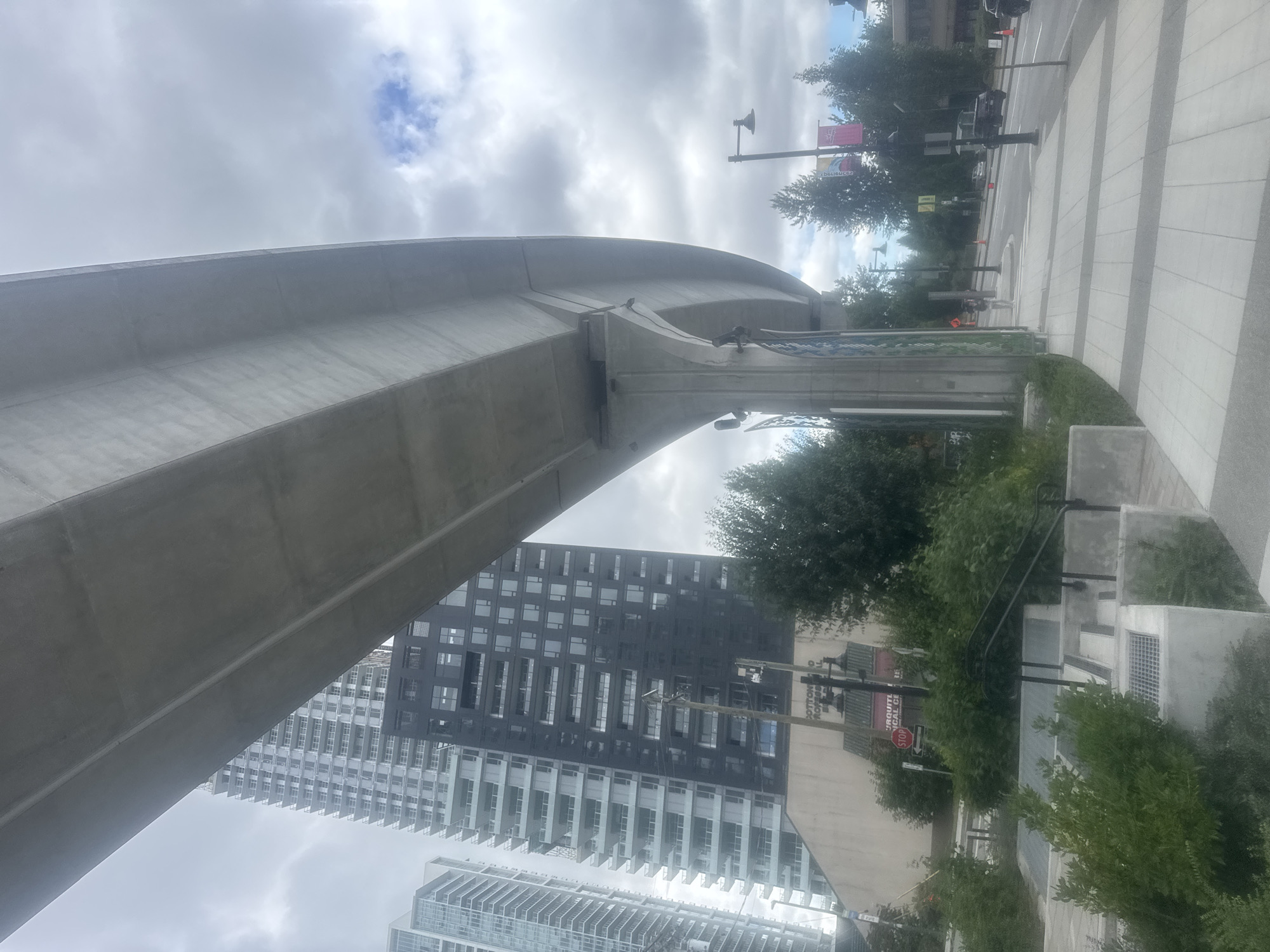 The image depicts an elevated transit line structure, likely an overpass or sky train, dominating the foreground and midground of the scene. The concrete structure curves through the frame, casting a shadow on a paved walkway and street below. In the background, a tall, modern high-rise building with a dark facade and numerous windows stands prominently. To the left of this building, another, lighter-colored high-rise is partially visible.
The environment appears to be urban, with trees and greenery interspersed along the street and sidewalk. Streetlights and traffic cones are visible on the road. A stop sign is present on the left side of the image, near a building with signage that appears to read "COTTONWOOD PROFESSIONAL CENTRE." Several cars are present on the road. The sky is overcast with clouds, suggesting a partly cloudy day.
Based on the presence of the "COTTONWOOD PROFESSIONAL CENTRE" signage, this location is identifiable as Coquitlam, British Columbia, Canada. The image captures an aspect of the urban infrastructure and cityscape of this area.