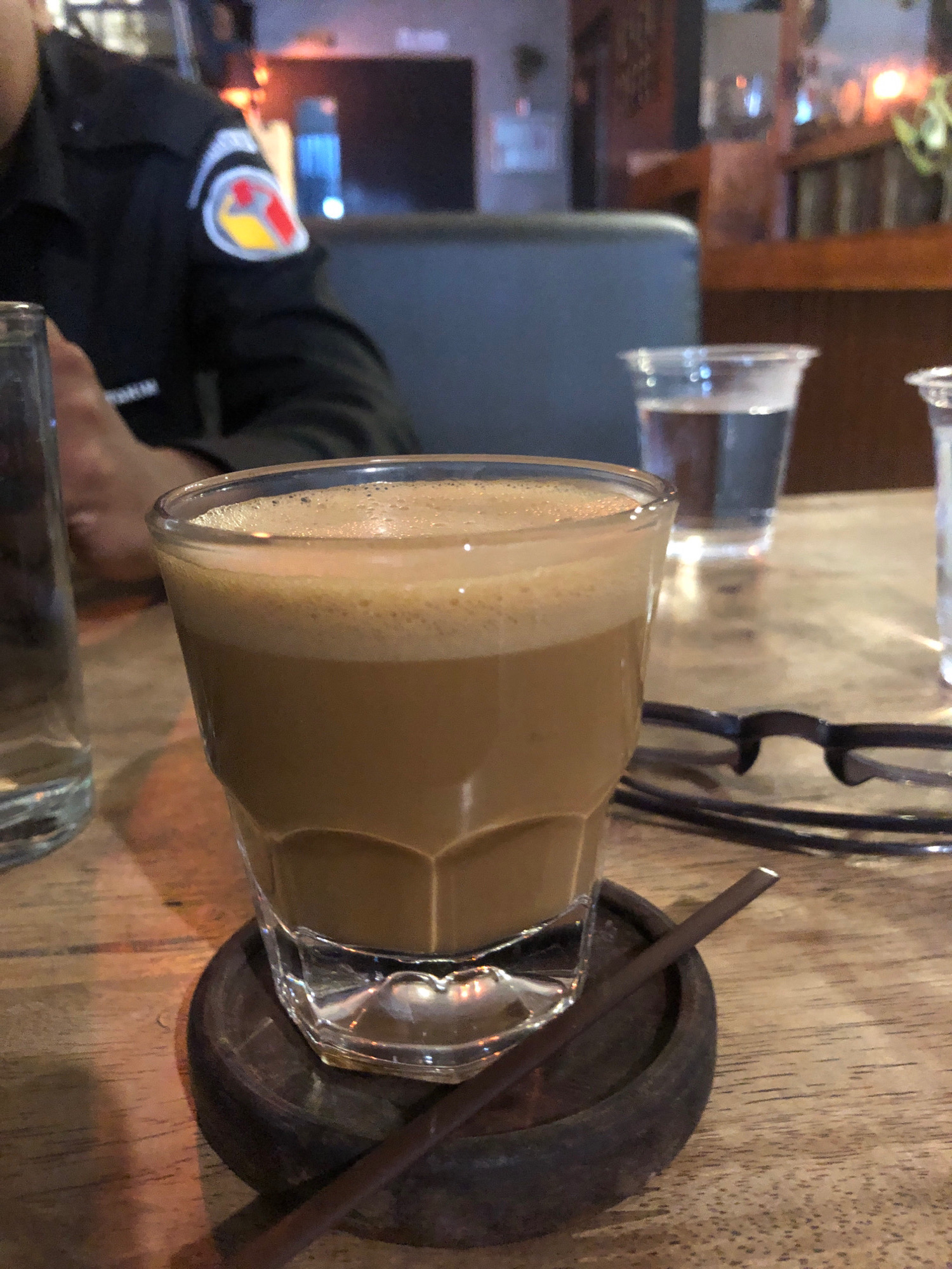 A close-up shot focuses on a glass of coffee with foam, placed on a wooden coaster on a wooden table. In the background, out of focus, are two plastic cups with beverages, and a person wearing a black uniform with a circular emblem on the sleeve. The environment appears to be an indoor establishment, possibly a cafe or restaurant. Given the context of Lhokseumawe, Indonesia, this could be a scene from a cafe in the city, a common spot for locals to gather and enjoy coffee.