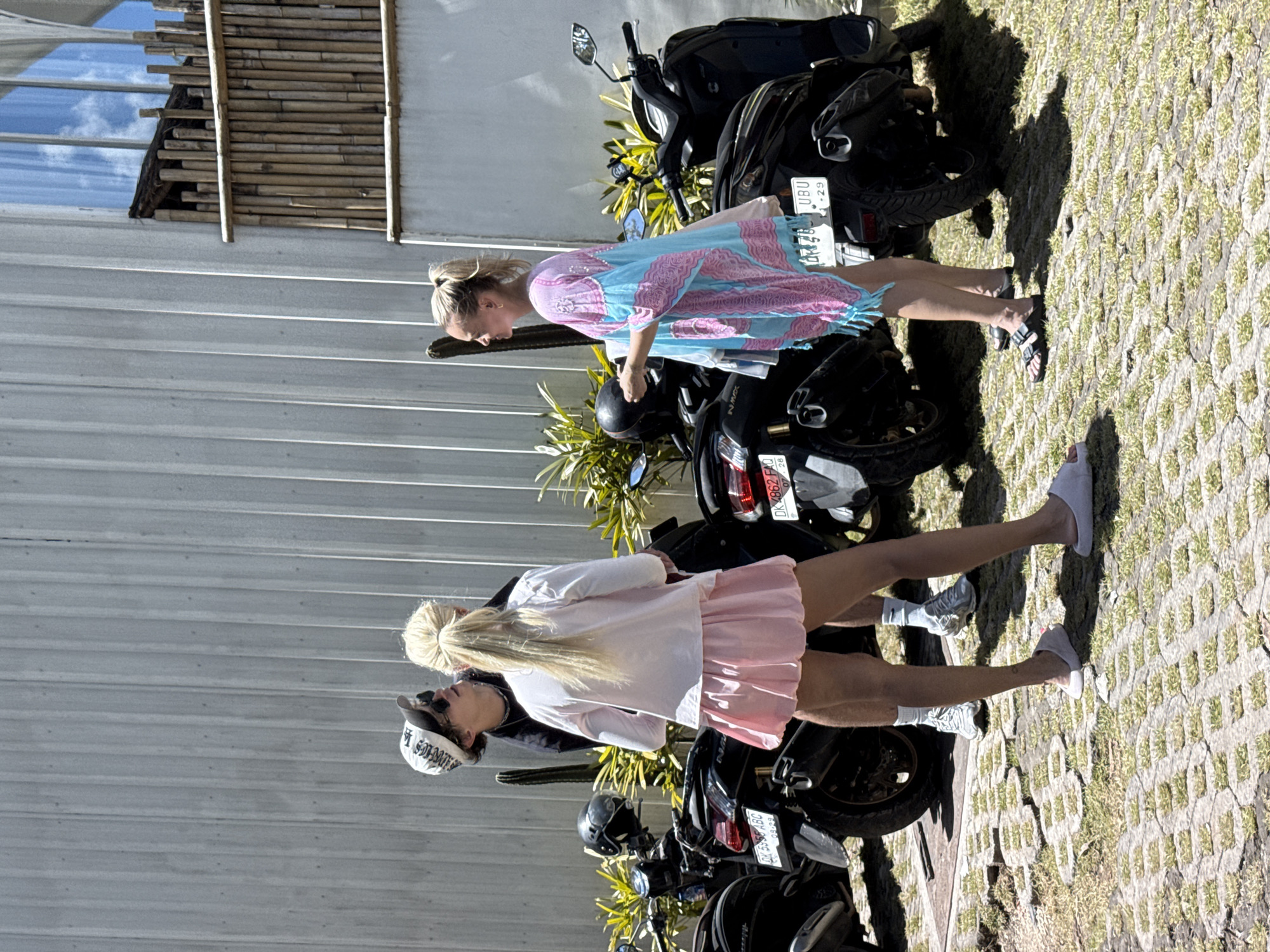 This bright outdoor photograph, rotated 90 degrees clockwise from its natural orientation, captures two women standing next to parked black scooters on a sunny day in Kuta Selatan, Indonesia. The ground is a light-colored permeable paving system with grass growing in the gaps, while a building with horizontal white siding and a decorative bamboo-slat structure forms the background.
In the foreground, a woman with long blonde hair tied in a high ponytail wears a white long-sleeved top, a pink pleated mini-skirt, white socks, and light-colored slip-on shoes. She also has sunglasses on her head and a white baseball cap with partially visible text "ST. SOME". She appears to be looking down towards the scooters.
Behind her, a second woman with blonde hair, also tied up, is seen in profile. She wears a loose-fitting blue and pink patterned caftan or cover-up and dark sandals. She stands near the rear of one of the scooters.
Several black scooters are parked, one displaying the model name "NMAX". Visible license plates include "DK 4862 FAQ 07-28" and "UBU -29". Strong shadows cast on the ground indicate bright sunlight, likely mid-day or afternoon, under a clear blue sky. A cactus-like plant is also visible near the scooters.