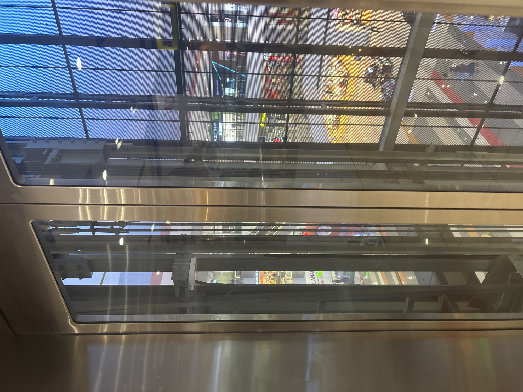 This image captures a view from inside a glass elevator in a multi-story shopping mall, likely located in Davao City, Philippines, given the prompt's context. The elevator interior, clad in polished stainless steel and featuring a glass wall, reflects the mall's interior. Through the glass, the mall's architecture is visible, including multiple levels with shops, escalators, and overhead lighting. The floors of the mall are a mix of colorful geometric patterns and white tiles. Several individuals can be seen walking on the different levels. The exterior of the mall is suggested by a large glass facade, possibly facing the sky, with a striped pattern resembling blinds or louvers. The overall scene depicts a modern, bustling commercial environment.