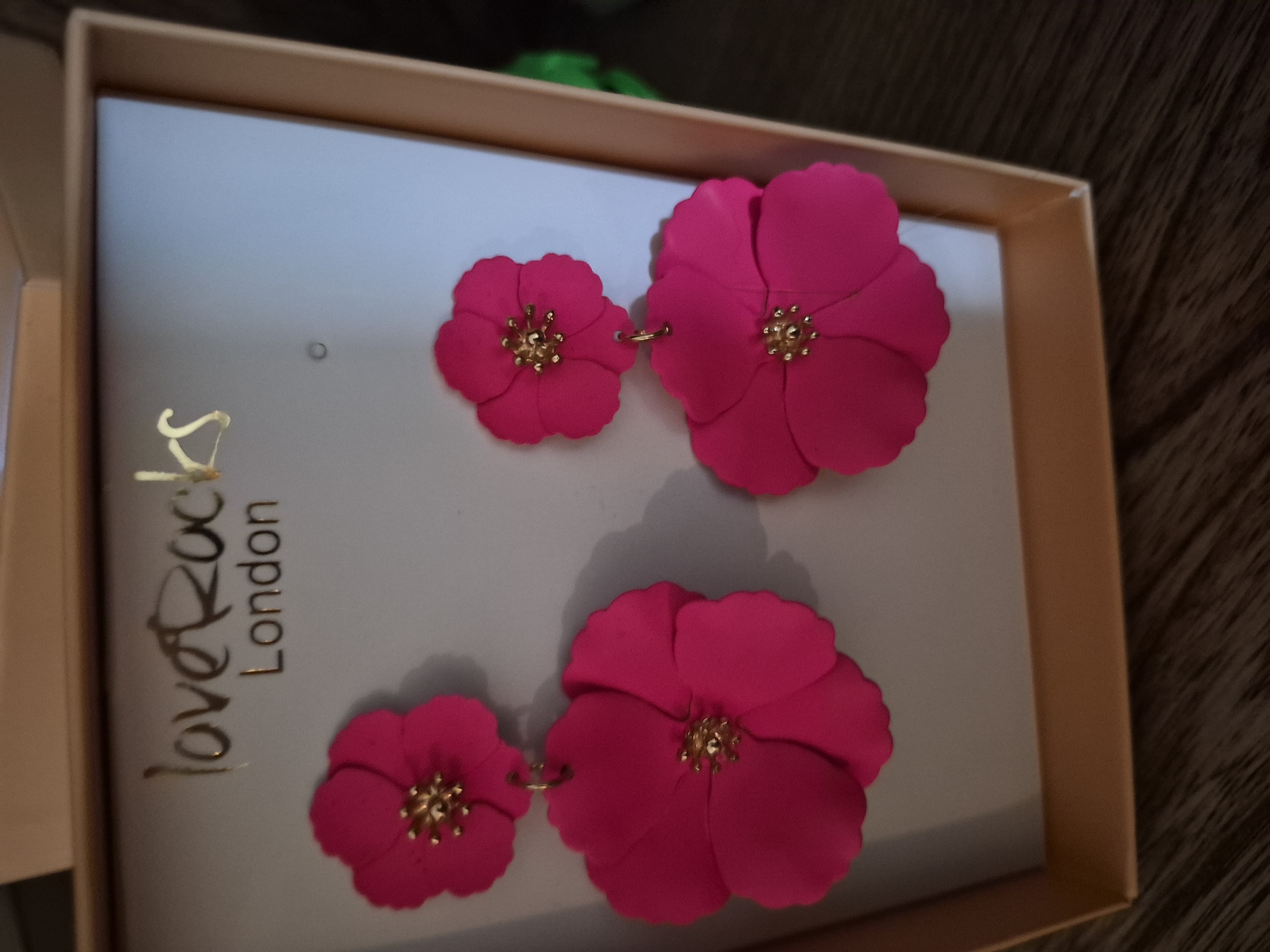 A pair of bright pink, double-layered, flower-shaped earrings are displayed in a gold-trimmed gift box. The earrings have gold accents in the center of each flower. The brand name "Love Rocks London" is printed in gold lettering on a light grey background above the earrings. The gift box is sitting on a dark wooden surface. The background is blurred.
