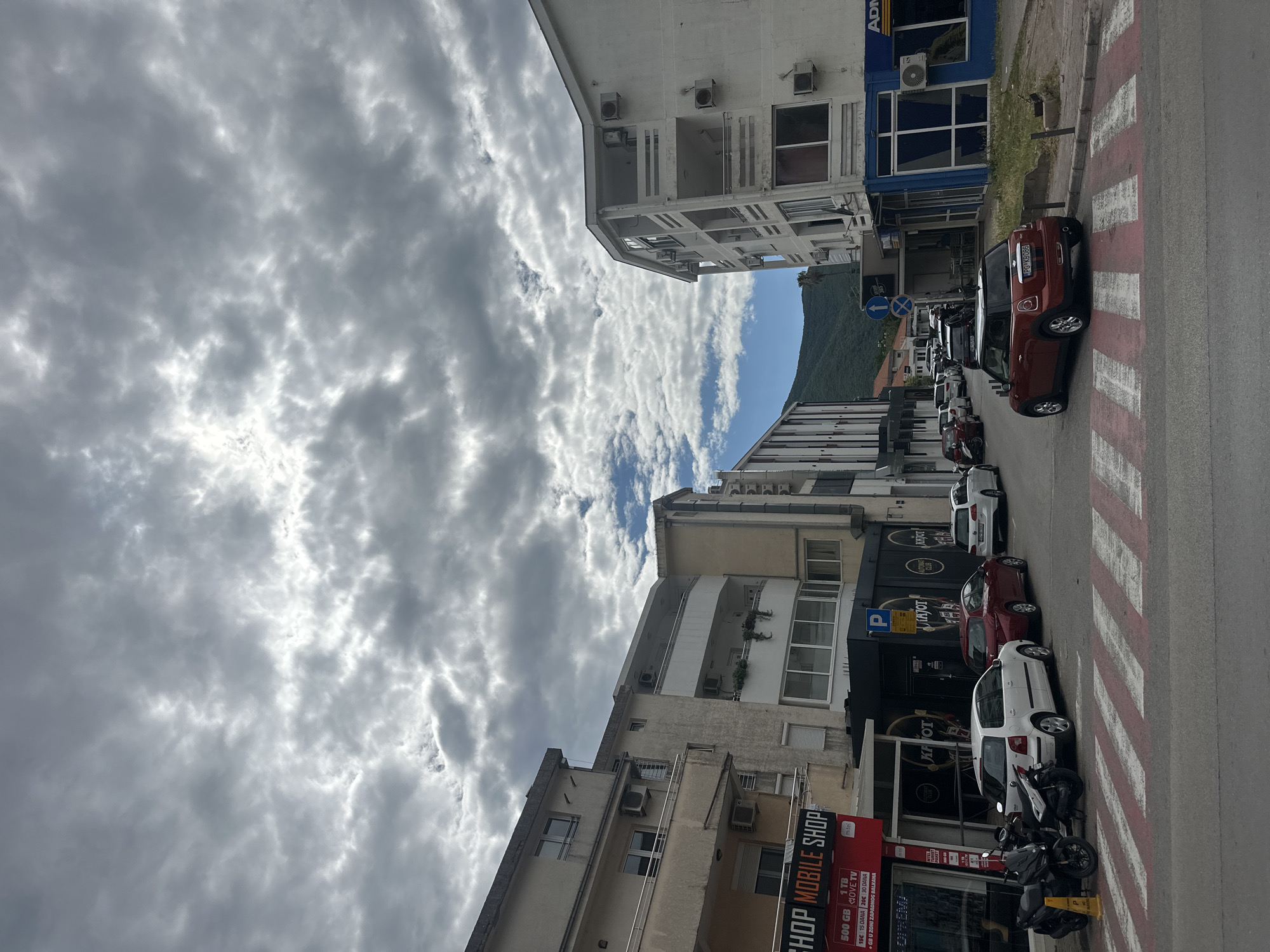 This image displays a daytime street scene in what appears to be a town, consistent with the provided location context of Budva, Montenegro. The view looks down a street flanked by multi-story buildings on both sides. A prominent green mountain range is visible in the background under a sky with significant cloud cover, interspersed with patches of blue.
The buildings lining the street house a mix of businesses on the ground floors and residential or office spaces above, characterized by balconies and numerous external air conditioning units. On the left side, a "SHOP MOBILE SHOP" sign is clearly visible, advertising "500 GB," "1 TB," and "MOVE TV," with pricing details like "15€ 15 DANA" (15 days) and "20€ 30 DANA" (30 days), and the additional service "+GB U ZONI ZAPADNOG BALKANA" (GB in the Western Balkans zone) and "OPREMA" (equipment). Further down, another building features a sign with "KAJOT CLUB" text. A building on the right has a blue facade with part of the text "ADM."
Along the right side of the street, several vehicles are parked diagonally next to a pedestrian crosswalk marked with red and white stripes. The closest parked car is a red Mini Cooper, bearing the Montenegrin license plate "PG KB086." Other parked vehicles include various cars, such as a white and a dark red one, and a couple of motorcycles. A blue "P" sign for parking is also visible.
The scene is static, with no people actively present. The time of day is daytime, indicated by the lighting, and the weather is partly cloudy.