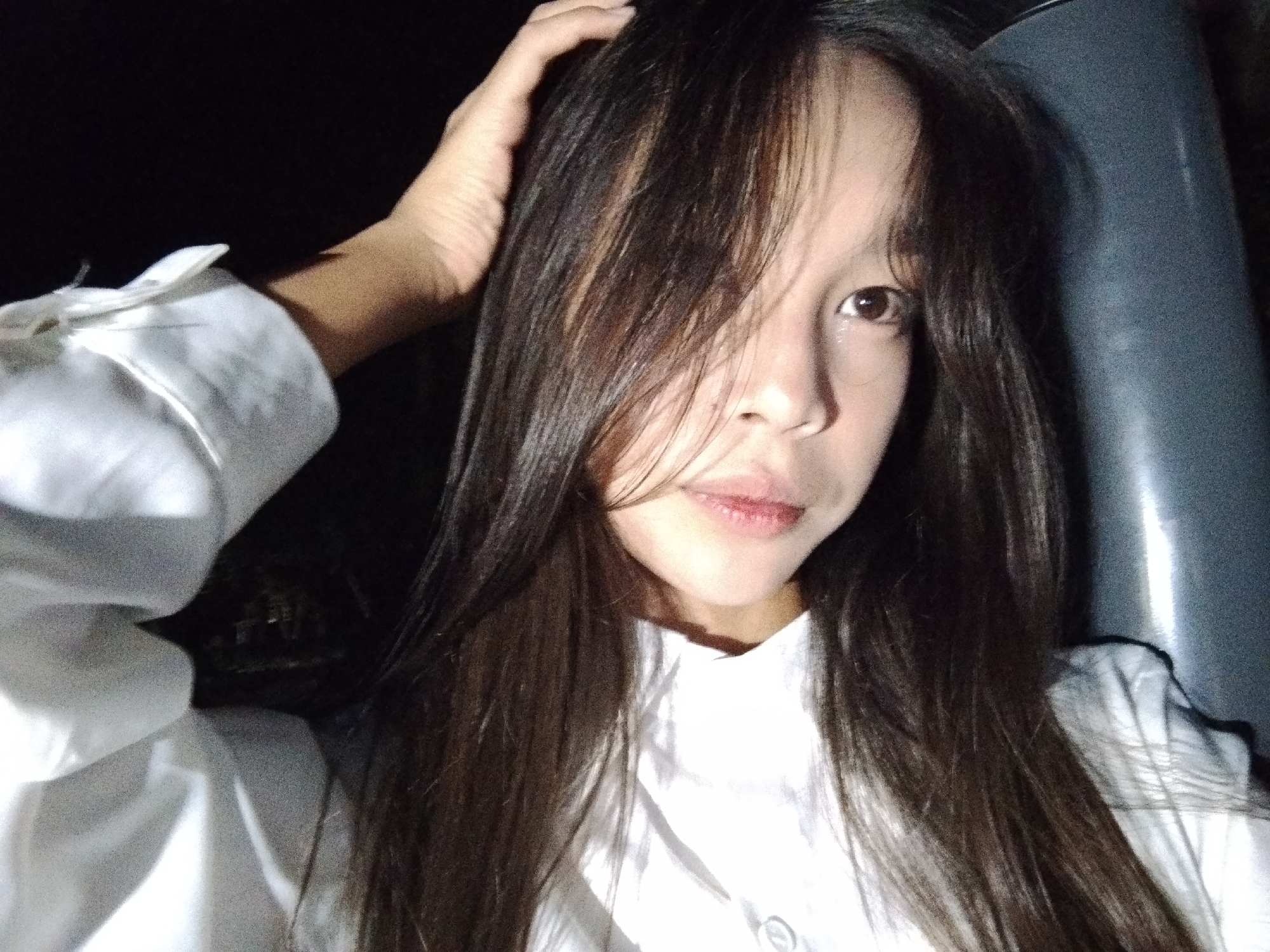 In a close-up, dimly lit photograph, a young woman is shown from the chest up. She has long, dark brown hair, some strands of which fall across her face, partially obscuring her right eye. Her left hand is raised and rests on the top of her head, with her fingers splayed. She is wearing a white collared shirt. The background is predominantly dark, with a vertical, grey cylindrical object visible on the right side of the frame. The lighting suggests the photo was taken at night or in a very dark indoor setting. There are no discernible landmarks or indicators to place this image within Bishkek City, Kyrgyzstan, or any other specific geographic location.