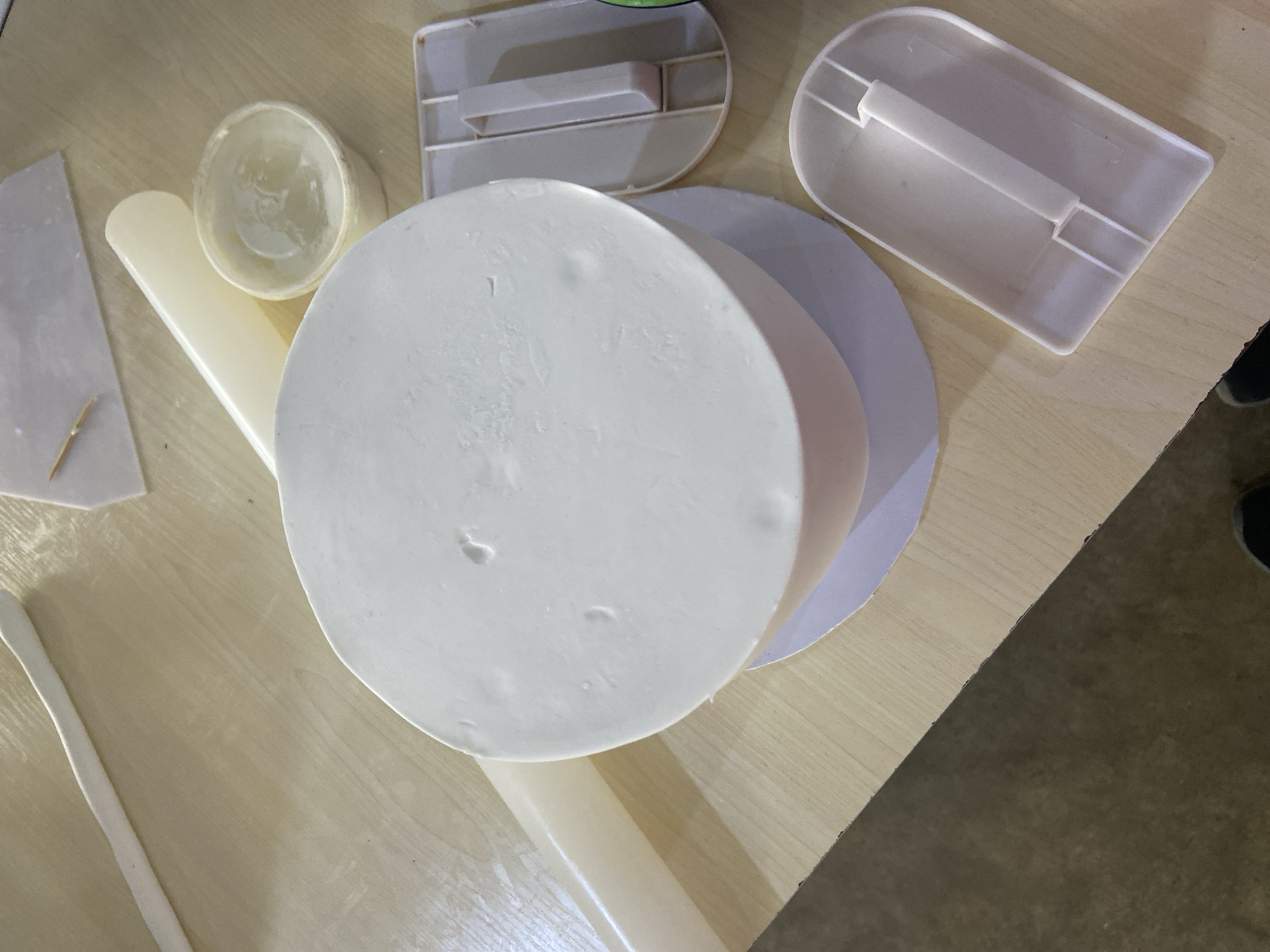 The image depicts a close-up, overhead view of a cake being decorated on a wooden table. A round cake, covered in white fondant, is the central subject. Various cake decorating tools are scattered around it, including a white rolling pin, a small plastic tub with a white substance, and two pale pink plastic scrapers. One scraper is positioned to the right of the cake, and another is below it. A small, thin wooden stick, possibly a toothpick, rests on a piece of translucent paper in the upper right corner. The background is a neutral-toned wooden surface. The scene is indoors, and based on the style of the table and general aesthetic, it could plausibly be located in a kitchen or workshop setting in Jos, Nigeria, where such cake decorating activities are common. No people are visible in the frame. The lighting suggests an ambient indoor light source.
