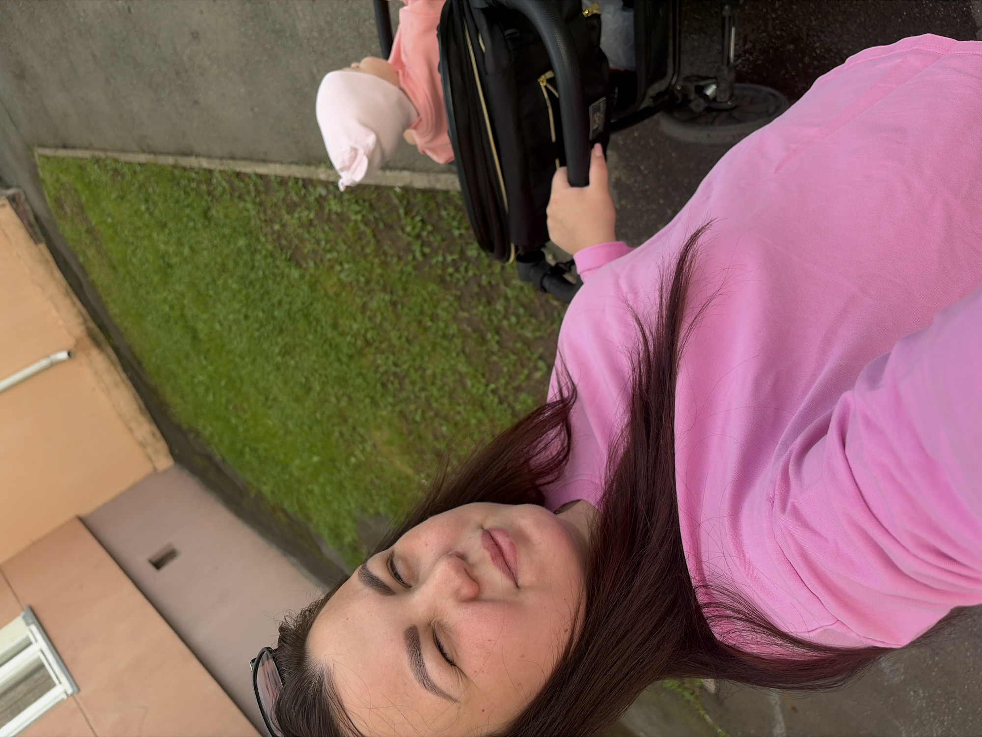 An overhead medium shot captures a woman with dark hair and a pink sweatshirt pushing a black stroller with a baby inside, who wears a pink bonnet. The woman is looking to her left with her eyes squinted. The background shows a building and a patch of green grass bordering a grey sidewalk. The scene is located in Izhevsk, Russia.