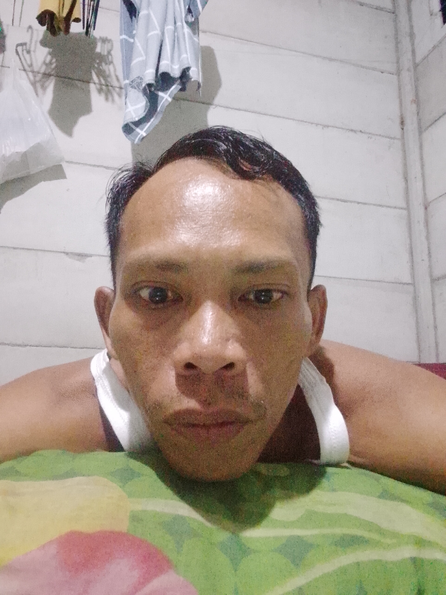 A close-up selfie shows a man with dark hair, brown eyes, and visible pores, lying on a patterned green and yellow pillow. He is wearing a white tank top. The background consists of a white wall, from which hang articles of clothing. The photo was taken indoors in Bukit Ketok, Indonesia.