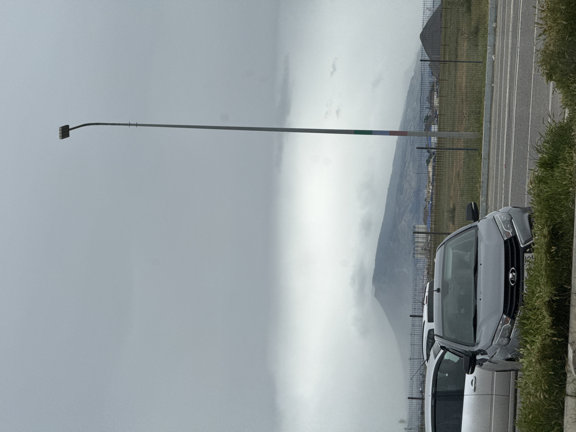This image captures an overcast outdoor scene in Kaspiysk, Russia, featuring a muted color palette and heavy cloud cover.
In the foreground to the right, two light-colored cars are parked on a paved surface. The closest vehicle is a Lada, identifiable by its front grille and the prominent Lada emblem. A partial view of its license plate shows "RUS" and digits including "17," confirming its Russian origin. Green grass and sparse vegetation line the edge of the parking area, which features white markings on the asphalt.
A tall, slender streetlight pole stands on the left side of the frame, topped with a modern, rectangular LED light fixture. Near the top of the pole, colored bands of green, white, and red are visible. A wire mesh fence stretches horizontally across the mid-ground, separating the paved area from the distant landscape.
The background features a range of mountains or hills, their lower sections enveloped by a thick band of mist or low-hanging clouds, contributing to the overall subdued and dim atmosphere of the daytime scene. No people are visible, and no specific event is taking place. The weather appears dull and hazy.