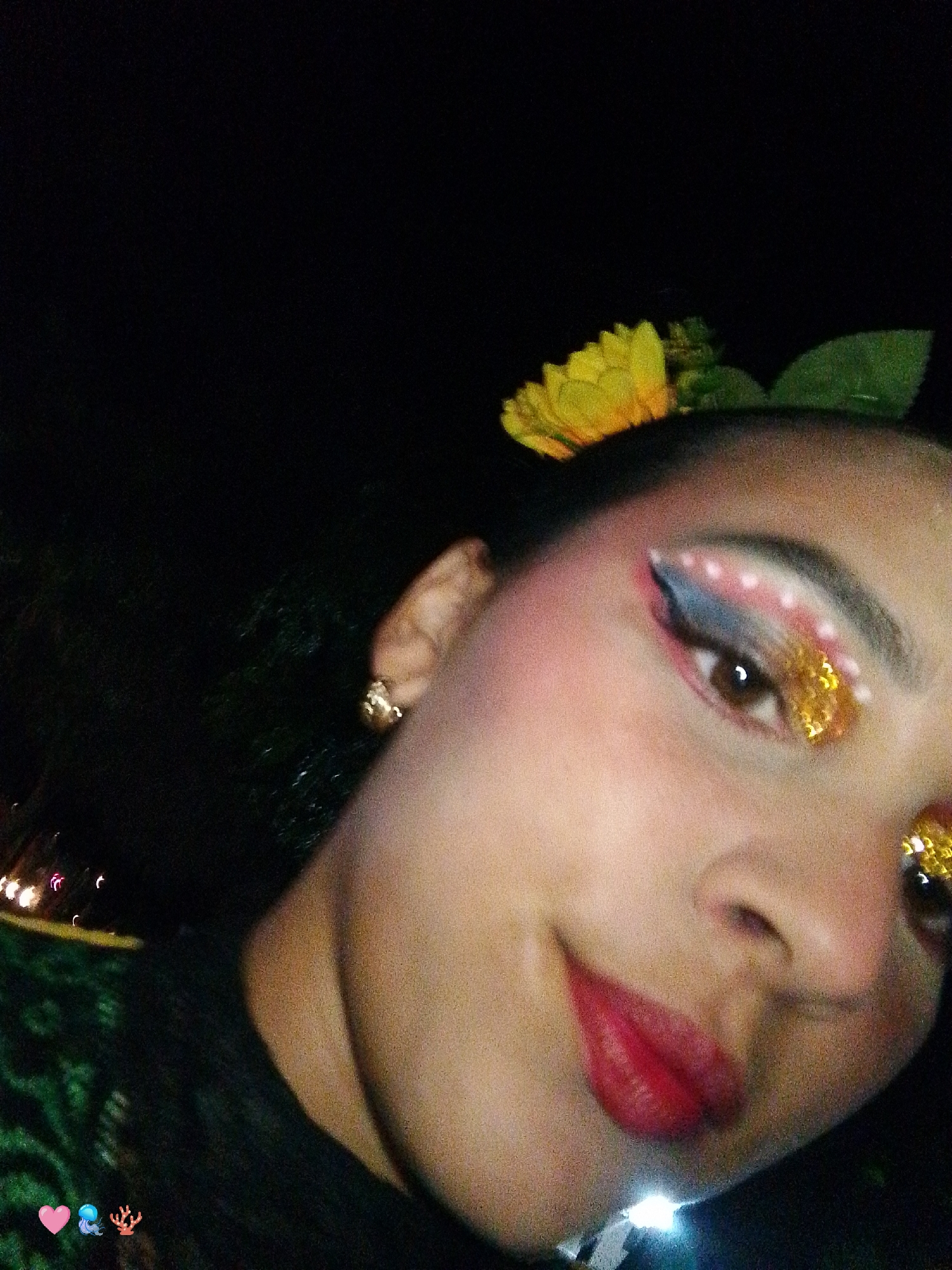 This is a low-light, close-up portrait, likely a selfie, of a young adult female in or around Playa El Agua, Venezuela. The individual's face occupies the majority of the frame, angled slightly, with her head tilted. Her hair, which appears dark, is adorned on the right side (viewer's left) with a bright yellow artificial flower and green leaves.
Her complexion is light to medium, with visible blush or contour on her right cheek. Both eyes feature elaborate makeup; the right eye (viewer's left) shows a blend of light blue and pink eyeshadow on the eyelid, accented by an arc of small white dots. The lower eyelid and crease are covered in chunky gold glitter. Her iris is brown, with a distinct light reflection. Her eyebrows are dark and well-defined. Her full lips are coated in a glossy, vibrant red lipstick, exhibiting a slight upward curve suggestive of a smile. A small, gold-toned stud earring is visible on her right earlobe (viewer's left).
Her attire includes a dark garment, possibly a top or dress, visible on her chest. A textured green fabric, potentially part of a costume or another clothing layer, is visible on the lower left side of the image. The background is predominantly dark and out of focus, indicating low ambient lighting conditions, consistent with evening or night. Blurred, warm-toned lights are visible in the distant background on the far left. In the lower left corner, three emojis – a pink heart, a blue jellyfish, and a red coral – are digitally overlaid on the image.
