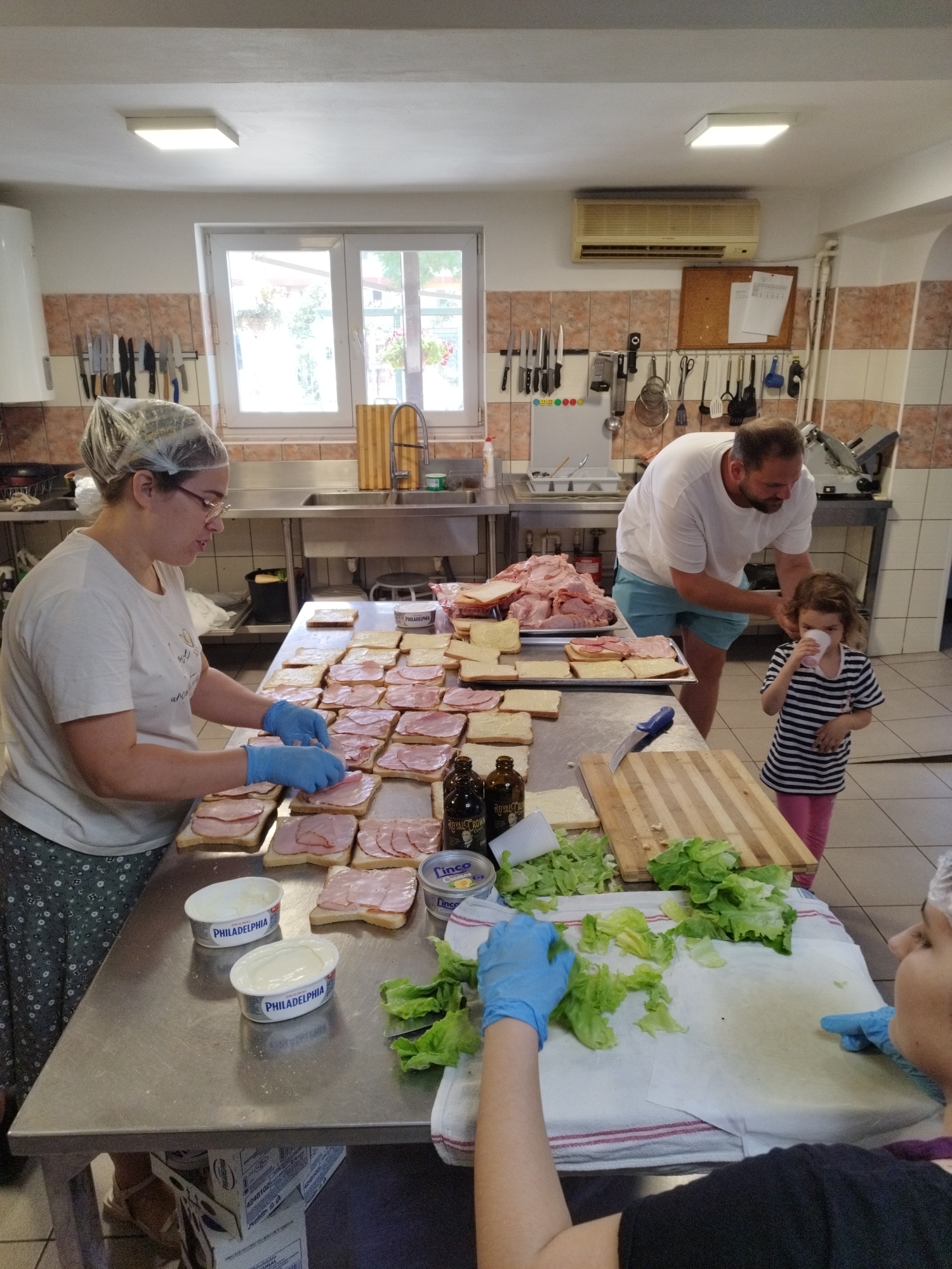 The image depicts a group of people preparing sandwiches in what appears to be a commercial kitchen. In the foreground, a woman wearing a hairnet and blue gloves is actively assembling sandwiches. She is placing ham on slices of bread that are already spread with a creamy substance, possibly cream cheese, indicated by two tubs of "Philadelphia" visible on the table. Adjacent to her, another person, mostly out of frame, is also wearing blue gloves and working with lettuce.
In the mid-ground, a man in a white t-shirt and blue shorts is standing next to a young girl in a striped shirt and pink pants. The girl is holding a cup to her mouth, and the man appears to be interacting with her. Behind them, there is a large pile of sliced meat, likely ham, and numerous prepared sandwich bases on trays.
The environment is a kitchen with stainless steel countertops and appliances. Knives are neatly arranged on a magnetic strip on the wall. A window is visible in the background, showing greenery outside. There is also an air conditioning unit mounted on the wall.
The overall scene suggests a large-scale food preparation event, possibly for an organization or a large gathering. The presence of commercial kitchen equipment and the organized assembly line for sandwiches indicate a functional and busy setting. The image does not provide specific visual cues to pinpoint the exact location within Bucharest, Romania, but the setting is consistent with a commercial or institutional kitchen.