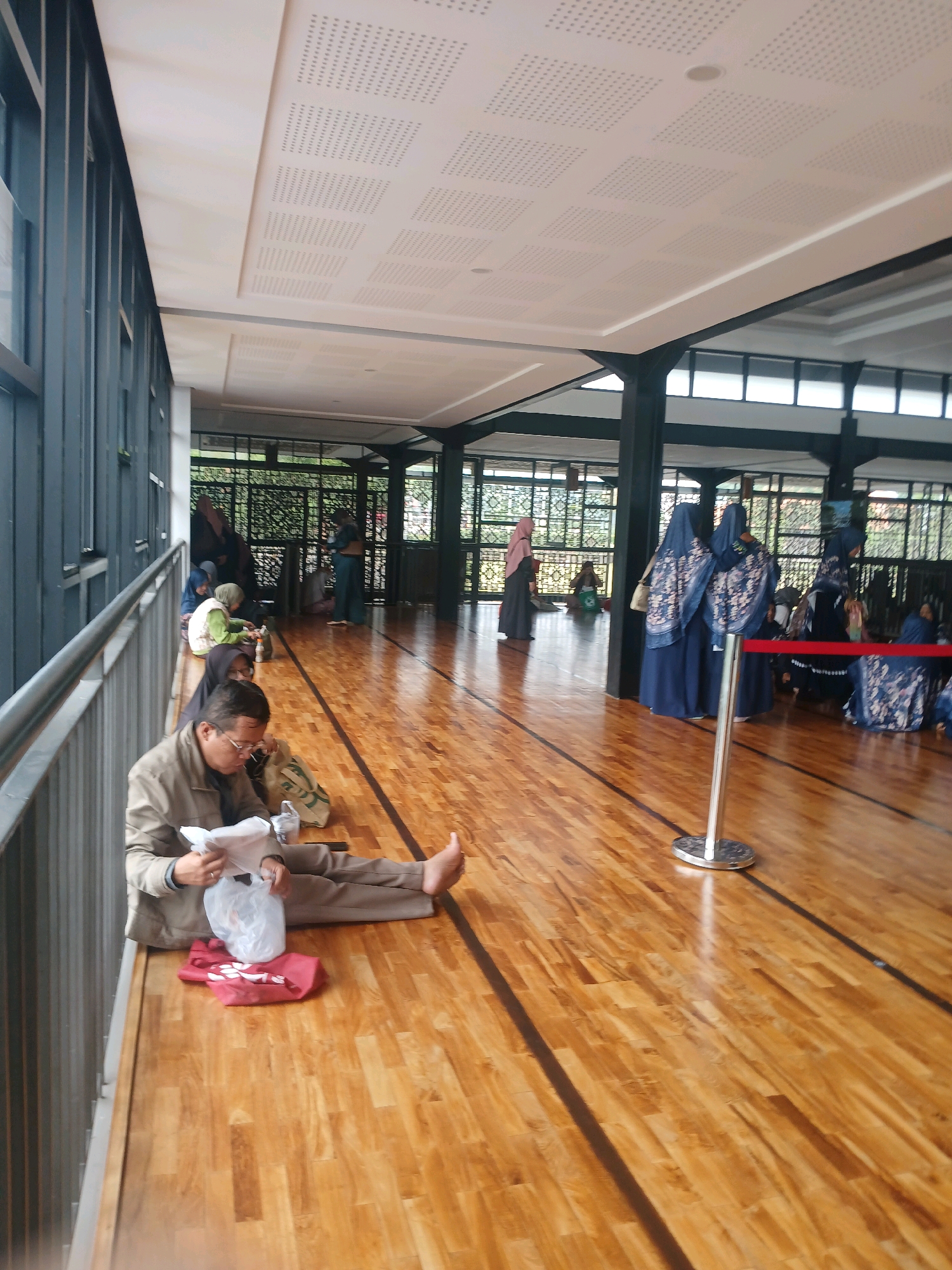 The image shows a group of people waiting in a covered outdoor area with a wooden floor. The area appears to be part of a larger building or complex, possibly a public space or a facility. The architecture includes dark pillars and a white ceiling with a grid pattern. There are large windows or openings along one side of the area, revealing greenery outside.
In the foreground, a man wearing glasses and a light brown jacket is sitting on the wooden floor, reading a document and holding a white plastic bag. He is barefoot and his legs are extended. Next to him is a red cloth item and a light-colored bag.
Further into the background, several other individuals are seated on the floor, some wearing hijabs and long garments in various colors, including blue, green, and pink. Some are engaged in conversations or looking at their phones. A red rope barrier with a silver stanchion is visible in the middle ground, suggesting a designated waiting or queuing area.
The scene suggests a communal waiting area, possibly at a religious site, a transit hub, or an event venue. The overall atmosphere is one of quiet waiting. The location is identified as Arjasari, Indonesia.