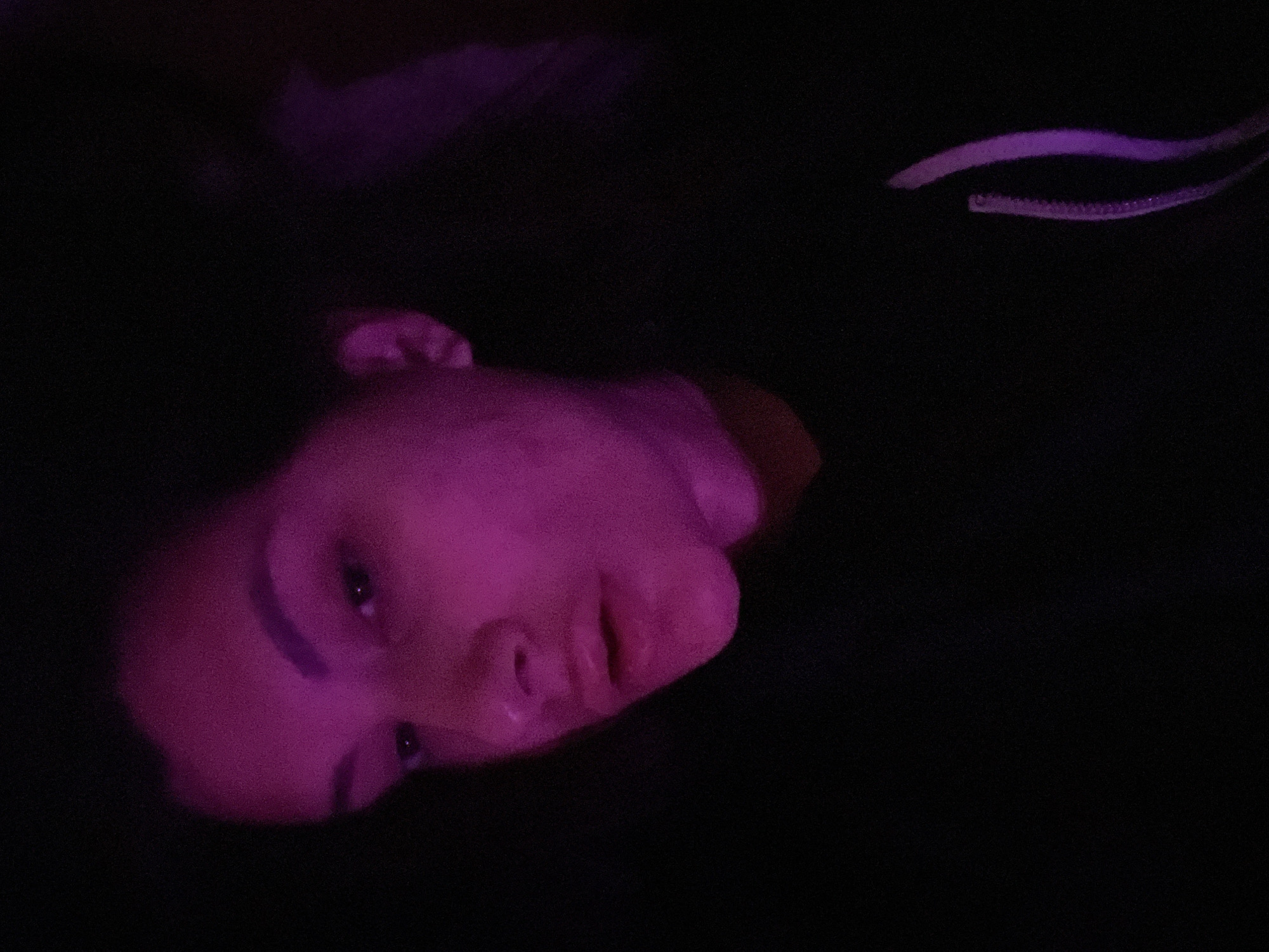 A close-up, dark image of a person's face illuminated with pink light. The person appears to be lying down, with their head tilted. The background is mostly black and indistinct, with possible hints of fabric. It's impossible to determine location from this image.