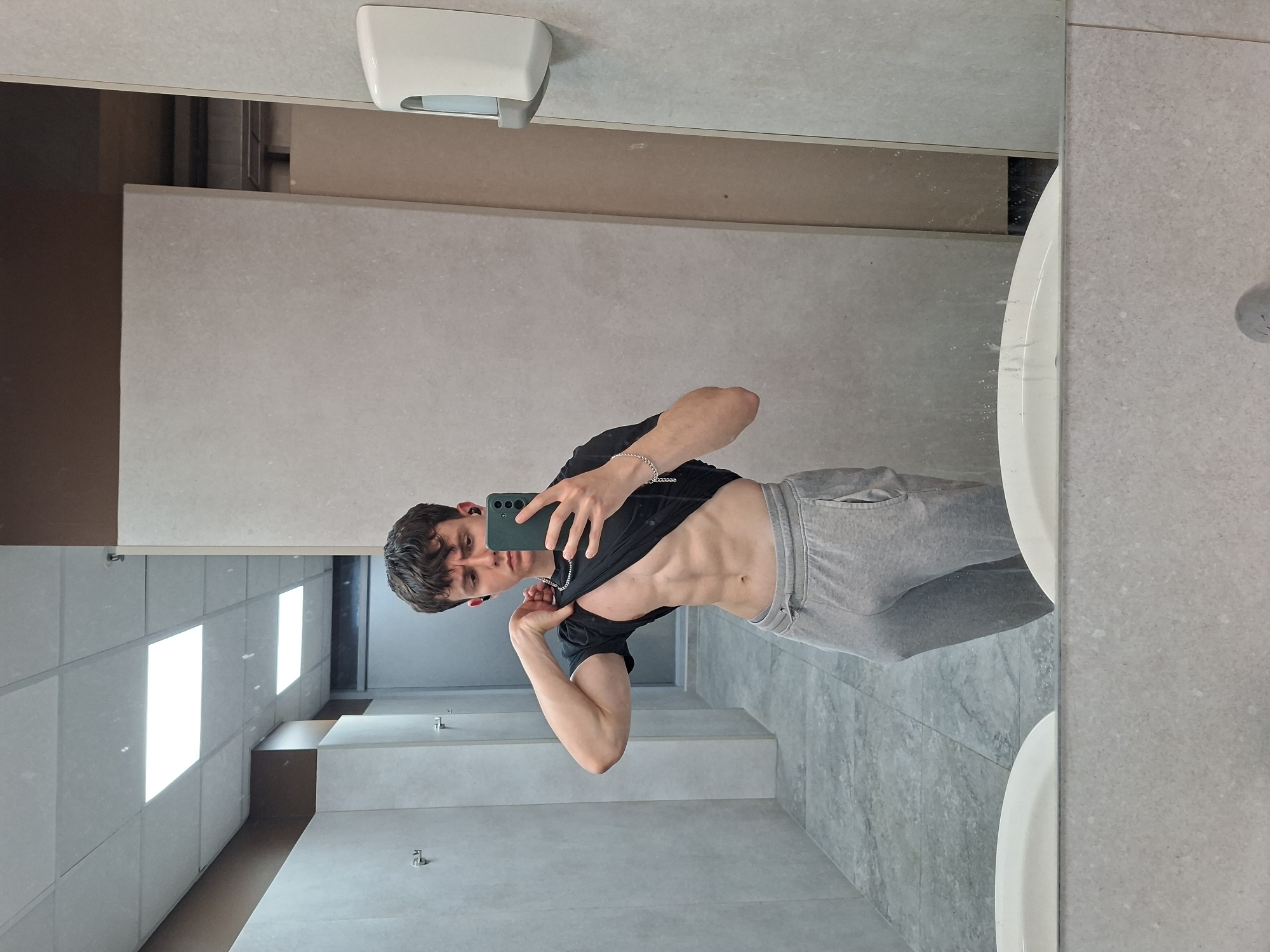 The image shows a young man taking a selfie in a bathroom. He is wearing grey sweatpants and a black t-shirt which he is holding up to reveal his abdominal muscles. He is holding a green smartphone. The bathroom has light grey tiled walls and floor, with a white sink and soap dispenser visible in the mirror. The lighting appears to be from recessed ceiling lights. The location is not discernible as Plovdiv, Bulgaria.