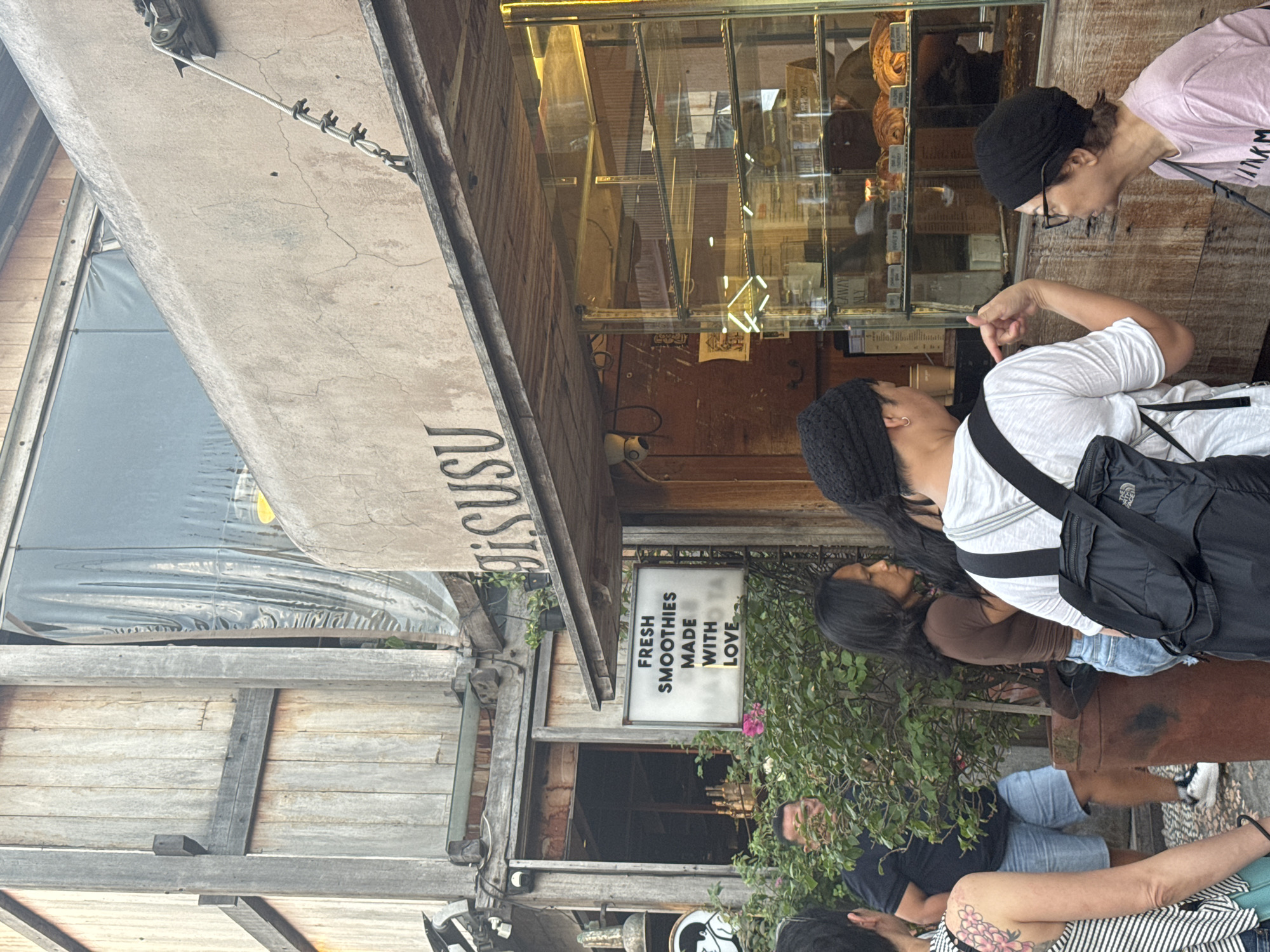 A group of people are gathered outside a food establishment with a sign that reads "GISUSU" and another sign that says "FRESH SMOOTHIES MADE WITH LOVE." The building features a rustic design with wooden elements and weathered concrete. A display case showcases baked goods. Some individuals are looking at the display or conversing. The scene appears to be in North Kuta, Indonesia, given the context.