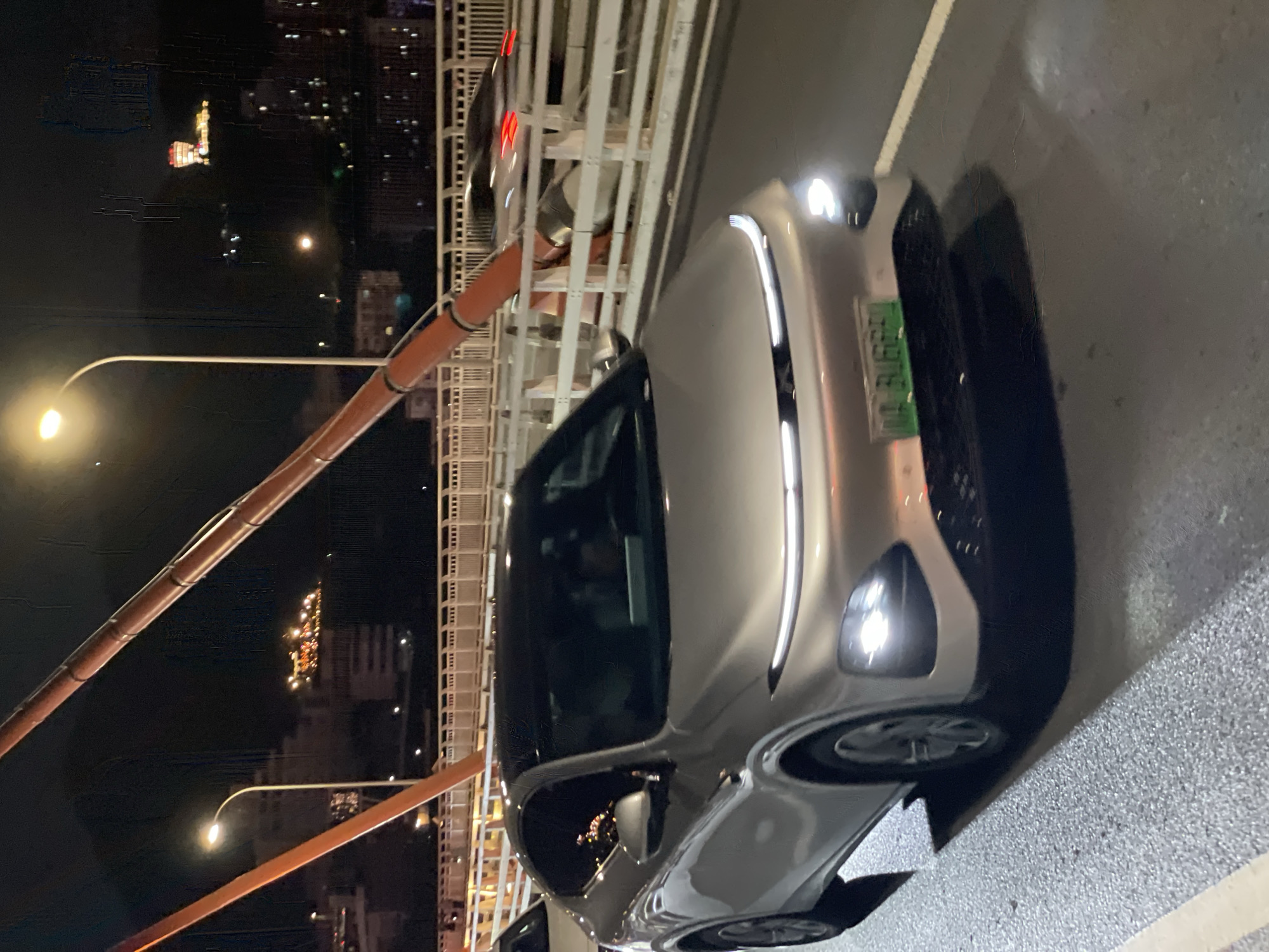 A silver, modern electric car, likely a crossover SUV, is driving on a bridge at night in Yuzhong District, China. The car has a distinctive LED light bar across the front and illuminated headlights. Its license plate is green with white characters. The bridge has orange-colored structural supports and a white railing. In the background, city lights are visible on the hillsides, indicating an urban environment. One building in the distance is lit up with colorful lights, appearing as a prominent landmark. The road surface is wet, suggesting recent rain. Another car is partially visible in front of the main subject, also driving on the bridge.