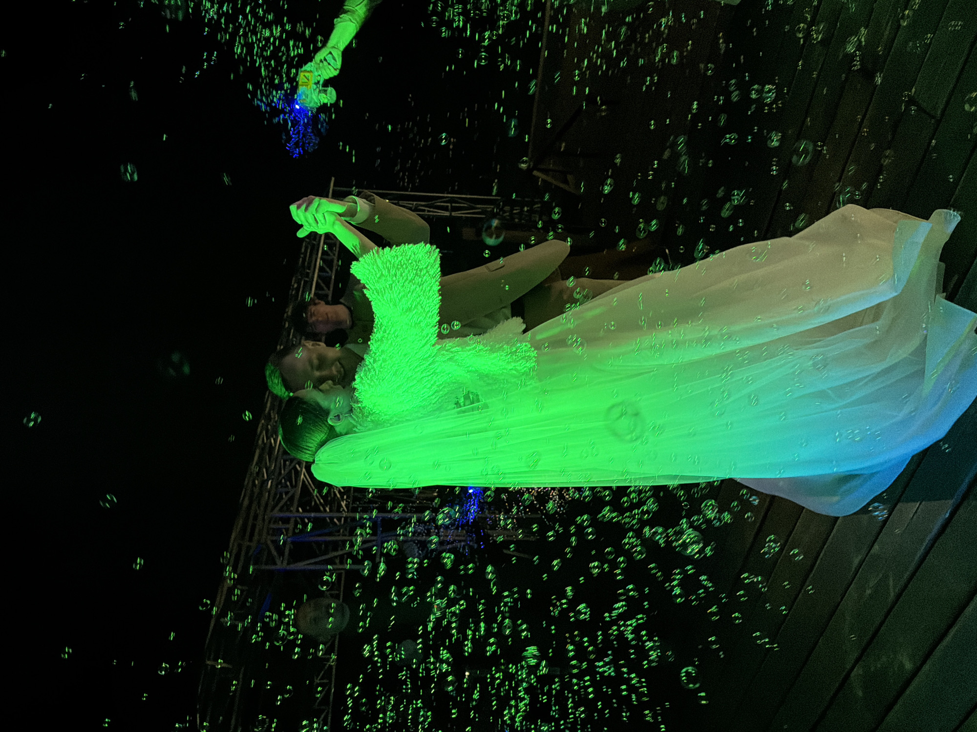 The image shows a couple dancing, likely at a wedding reception, at night. The scene is illuminated with green and blue lighting. Bubbles fill the air around them, possibly created by a bubble machine held by a person partially visible in the upper left. The woman is wearing a white gown, and the man is wearing a suit. A metal structure is visible in the background. The floor is made of dark wooden planks. The location, Chaikovsky, Russia, is based on prior knowledge and cannot be determined from the image alone.