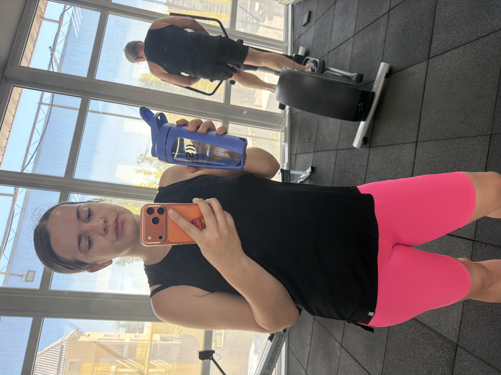 The image captures a mirror selfie taken by a woman inside a gym or fitness center, located in Sochi, Russia. The woman, positioned in the foreground and oriented sideways, is wearing a black sleeveless top and bright pink bike shorts. She holds a purple water bottle in her right hand, which has "((S))" and "ML200" (visible as "200ML" in reflection) text. In her left hand, she holds an orange smartphone with a triple-lens camera setup, used to take the selfie. Her hair is pulled back, and she has a slight pout.
In the background, reflected in the large windows, a man with light hair is visible from behind, wearing a black tank top and black shorts. He appears to be exercising on a piece of gym equipment, possibly a rowing machine or elliptical. The gym features dark gray tiled flooring.
Through the large windows, a bright, sunny outdoor scene is visible, suggesting it is daytime. A clear blue sky, some residential buildings, and what might be awnings or shade structures can be seen outside. The overall atmosphere is bright and active, typical of a gym environment.