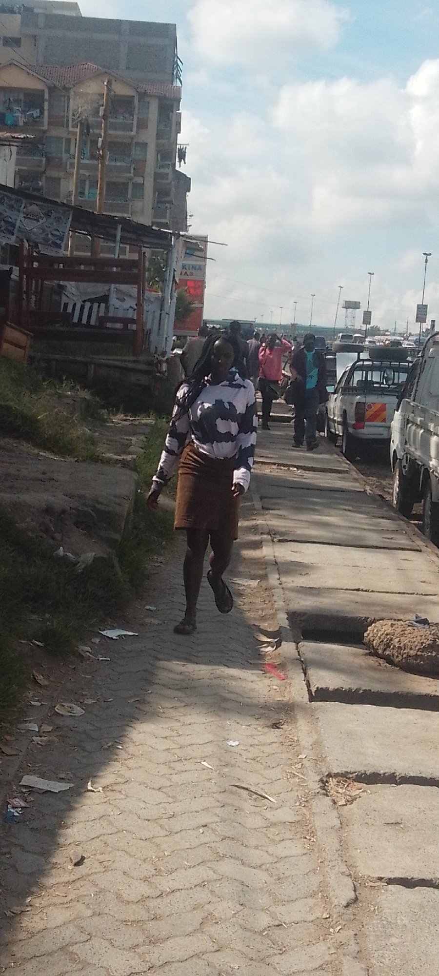 A woman walks on a concrete pavement in Nairobi, Kenya. She is wearing a striped long-sleeved shirt and a brown skirt. Other people are visible in the background, along with vehicles parked on the right side of the pavement. A multi-story building is situated on the left side of the image. The sky is partially cloudy.