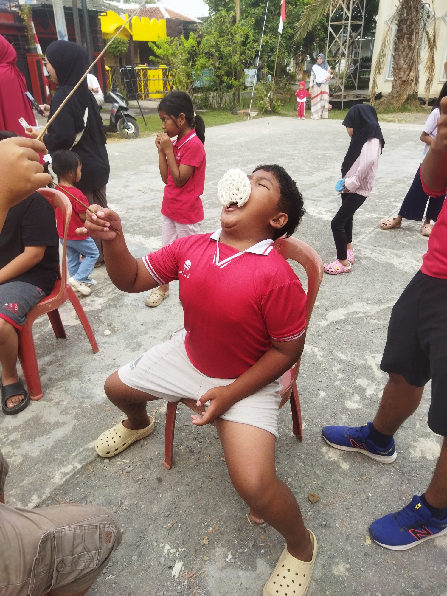 In a street setting in Banjar, Indonesia, a group of children participates in a cracker-eating contest. The main subject, a young boy in a red shirt and white shorts, sits on a red chair, attempting to eat a cracker suspended from a string by biting into it. His eyes are closed, and his mouth is open. Other children are visible in the background, some observing and others waiting their turn. A woman in a black hijab stands to the left, holding a string. The ground is paved with concrete, and trees and buildings are visible in the background, suggesting an outdoor event.