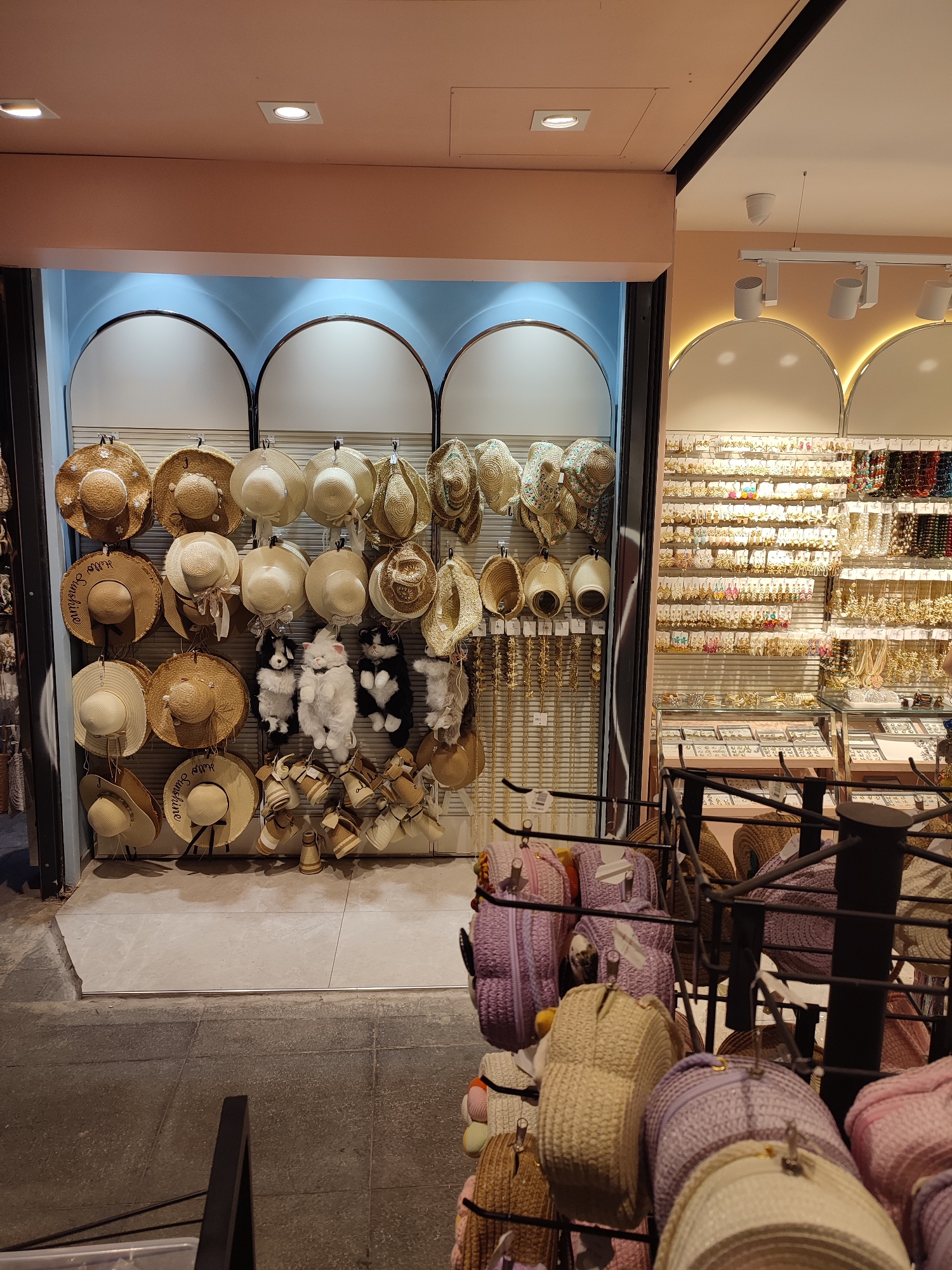 The image depicts the interior of a retail store, possibly a boutique or market stall, in Kadıköy, Turkey. The store features a wide array of accessories, primarily hats and jewelry.
The foreground displays a rack filled with woven bags, predominantly in shades of purple and beige. These bags are made of straw or a similar natural fiber and exhibit various textures and designs, including some with zipper closures.
Further back, a prominent display showcases a large collection of straw hats of different sizes and styles. Some hats are plain, while others are decorated with ribbons, embellishments, or written phrases like "Hello Sunshine." Interspersed with the hats are plush toys resembling cats and dogs.
To the right, an extensive wall display is dedicated to jewelry. Countless rows of earrings, necklaces, and bracelets are neatly arranged on hooks and shelves. The jewelry items are varied in color and design, with a notable presence of gold-toned pieces and colorful beads.
The store's interior features a clean and organized presentation, with neutral-colored walls and lighting that illuminates the merchandise. The overall impression is one of a well-stocked shop offering fashion accessories.