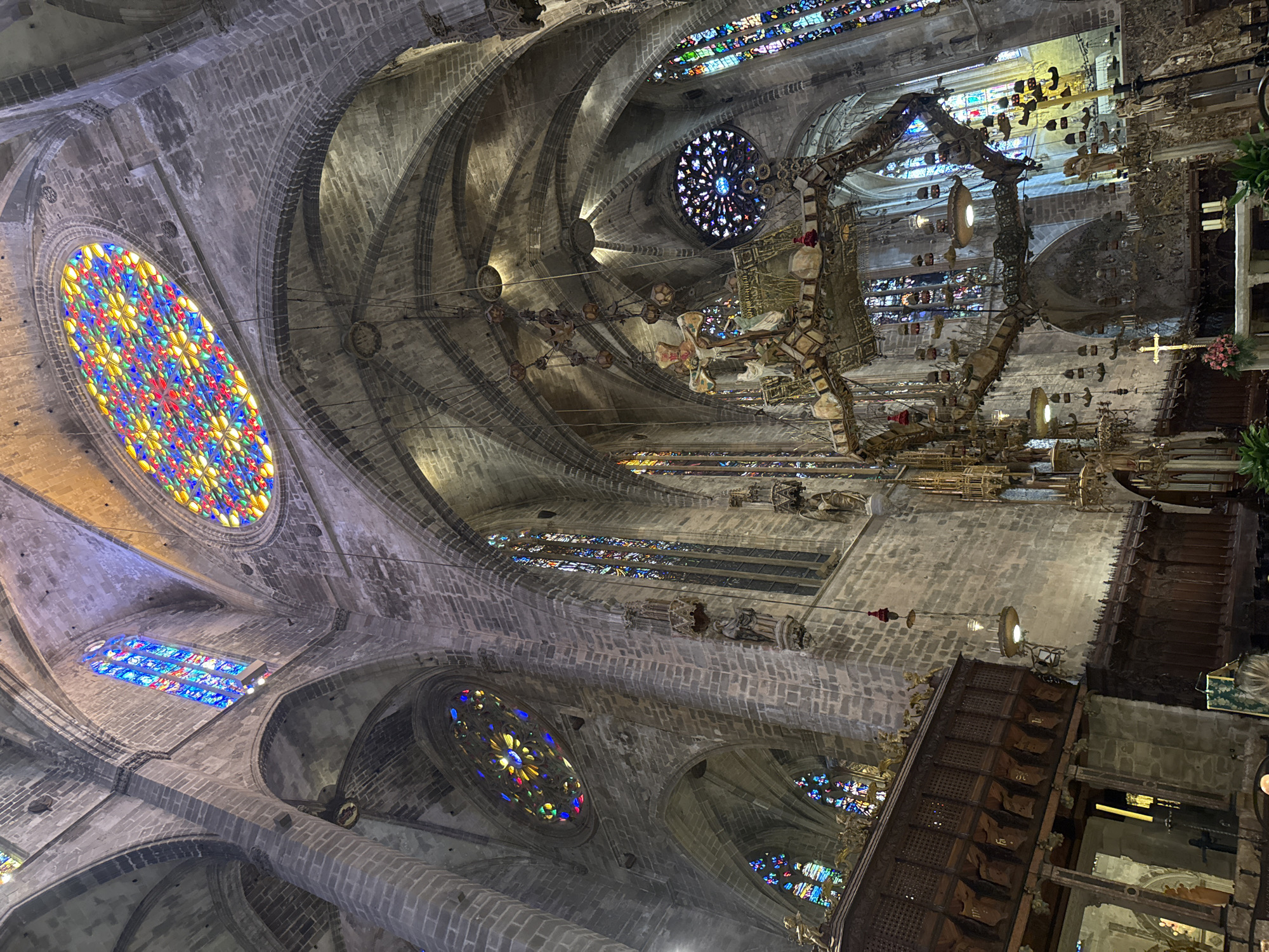This image shows the interior of the Palma Cathedral in Palma de Mallorca, Spain. The cathedral is a Gothic Roman Catholic cathedral. In the image, we can see high vaulted ceilings made of stone, supported by large pillars. There are stained-glass windows of various shapes and sizes throughout the cathedral, casting colorful light into the space. One prominent circular stained-glass window, also known as a rose window, is visible in the upper portion of the image. Suspended from the ceiling is a large, ornate chandelier. The lower part of the image shows wooden seating, likely choir stalls, and parts of an altar area with candles. The overall impression is one of grandeur and historical significance, characteristic of a large European cathedral.