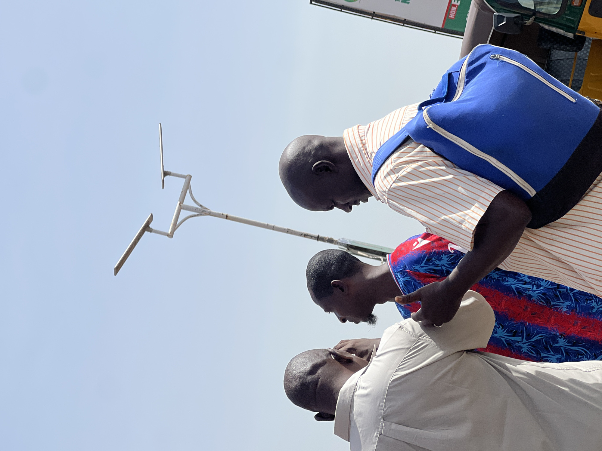 This outdoor image, captured in Jos, Nigeria, presents a low-angle view of three men under a clear, bright blue sky on what appears to be a sunny day.
The foreground features the top of a bald man's head, partially obscured, wearing a light-colored, possibly beige or light grey, long-sleeved shirt. Adjacent to him, a second man with a short beard is visible, wearing a vibrant shirt with red, blue, and black patterns. He appears to be leaning over or reaching towards the first man's head with his right hand, suggesting interaction or conversation between them. Further back and to the right, a third bald man is seen from the side, wearing a white and orange striped short-sleeved shirt. He carries a distinct royal blue backpack with white zippers and straps, and is looking downwards, seemingly engaged with the others.
In the upper left portion of the frame, a simple white metal pole structure with two horizontal bars extends upwards, possibly a street light fixture or sign support. To the upper right, a green sign with partial white text, "HON. E" and possibly "EN" or "GN", is visible. Next to the sign, parts of a yellow and green vehicle, consistent with a Keke Napep (auto rickshaw), can be identified, providing a contextual clue for the Nigerian setting.