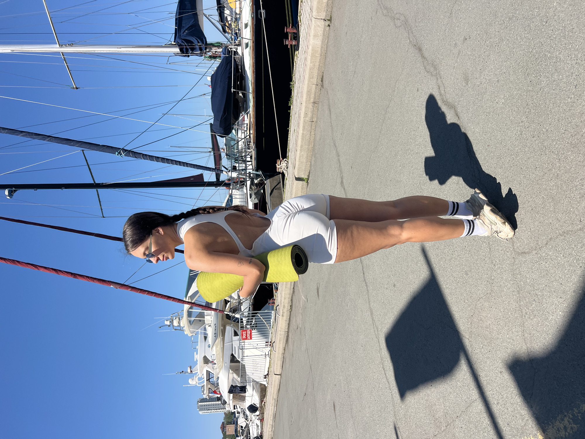 A woman wearing a white jumpsuit and striped socks stands on a gray paved surface, holding a rolled-up green yoga mat. Her hair is braided and she wears sunglasses. To the left, a large sailboat with masts and rigging is visible, along with other boats docked in a harbor. The location appears to be a marina in Vladivostok, Russia, as indicated by the Cyrillic signage and the cityscape in the background. The sky is blue, and the sun casts a shadow of a flag onto the pavement.