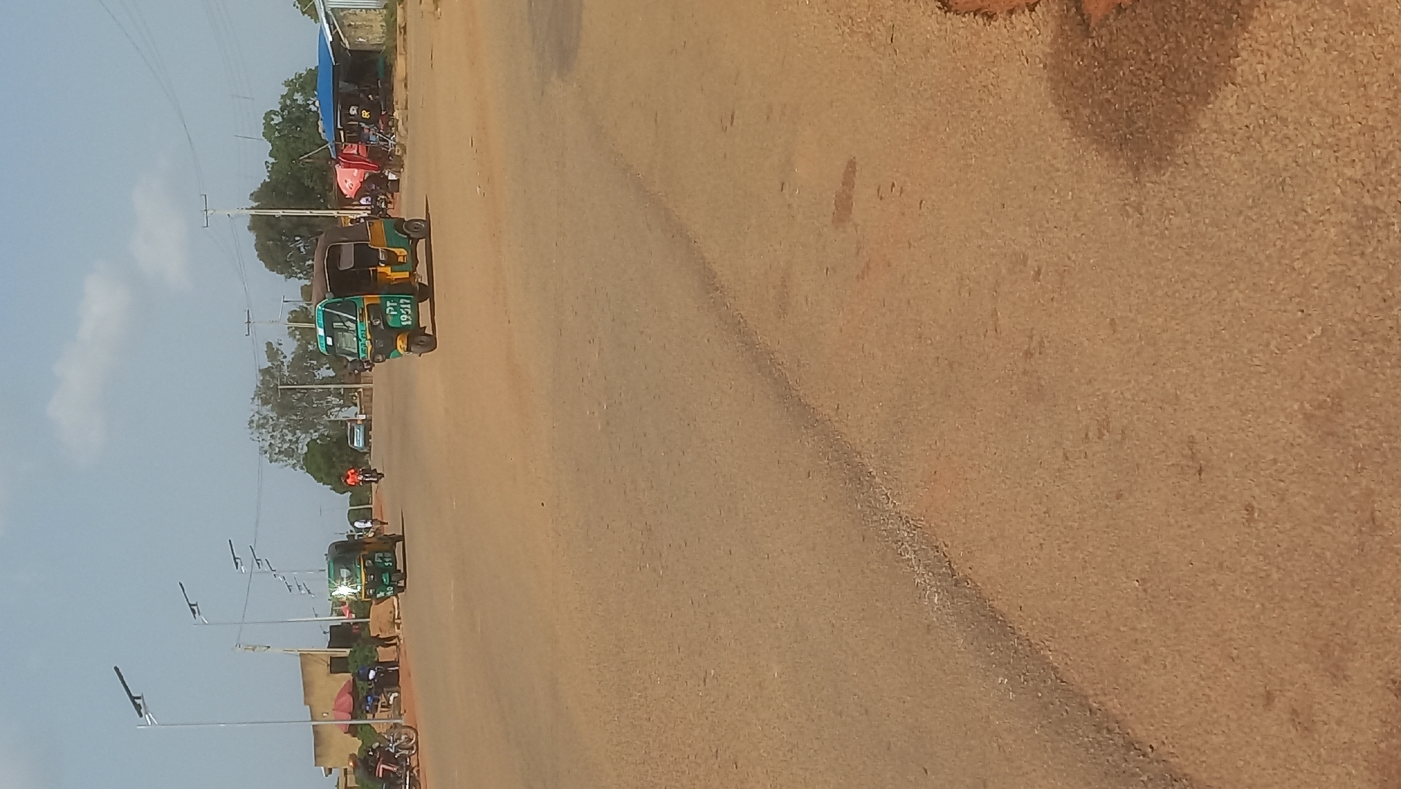 This media file depicts a street scene in Gura Top, Nigeria. The image is oriented vertically, with the ground filling the right side and the sky on the left. A dusty, reddish-brown road dominates the lower portion of the frame, with a darker, possibly paved, lane visible on the left side of the road. Several auto-rickshaws, also known as "keke napep" or "okada," are parked or driving along the road. One auto-rickshaw in the foreground is green and yellow, with the license plate "PT 19517" clearly visible. Other auto-rickshaws and motorcycles are scattered further down the road.
On the left side of the road, there are trees and some structures, including a building with a blue roof and what appears to be a market stall with red and white umbrellas. Lampposts are also visible along the side of the road. The sky is a pale blue with a few wispy clouds, suggesting a clear day. The lighting indicates it is daytime, likely mid-morning or afternoon. There are no people prominently featured in the foreground, but figures can be seen near the vehicles and stalls further down the road, suggesting activity and people going about their daily routines. The overall impression is of a bustling, local street in a Nigerian town. The reddish-brown earth and the types of vehicles are characteristic of many parts of Nigeria.