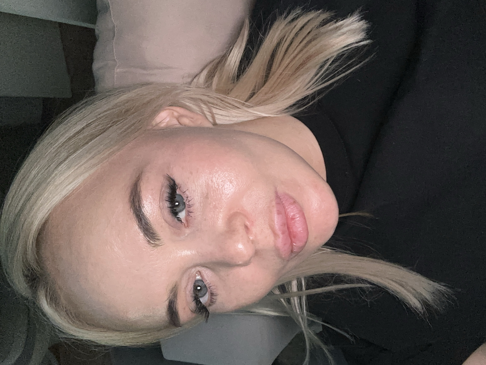 The image depicts a close-up of a woman's face. She has fair skin, light blonde hair, and light-colored eyes. She is wearing dark clothing. The background is out of focus, but includes a light-colored pillow. The image is rotated. The location in Высокий, Russia, is unknown.