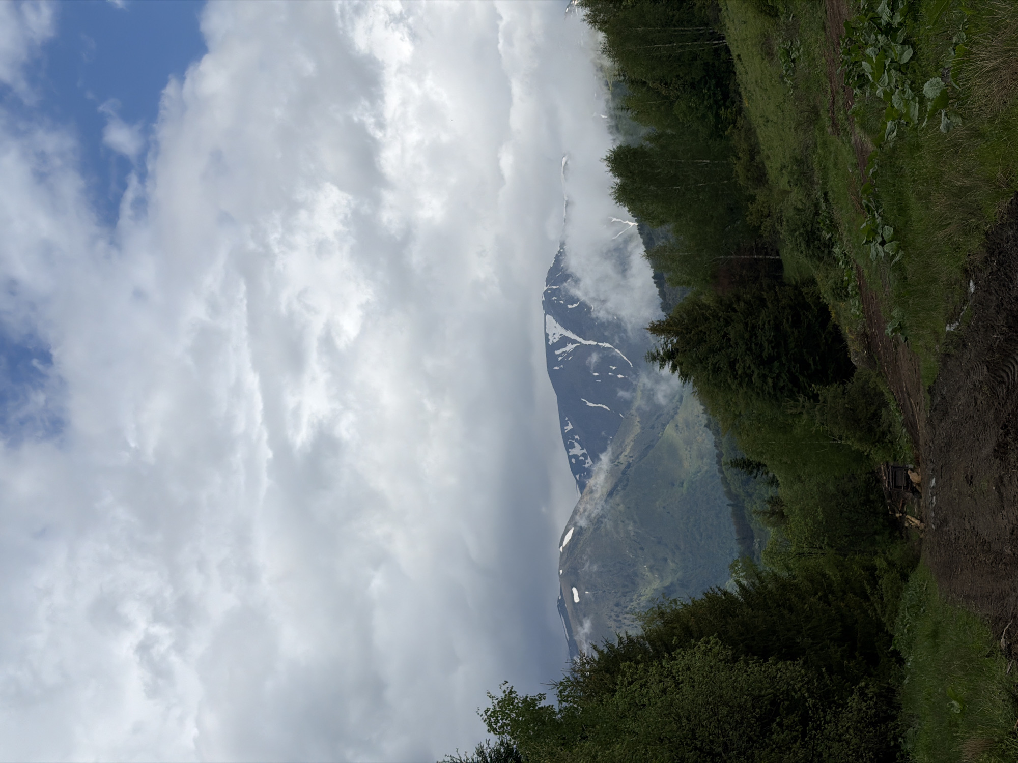 The image depicts a mountain range partially obscured by clouds, adjacent to a hillside path. Snow patches are visible on the mountain's slopes. The sky exhibits both blue patches and dense cloud cover. A dirt path with greenery borders the mountain view. The location is likely near Рилски манастир, Bulgaria, given the mountainous terrain and vegetation typical of the region.