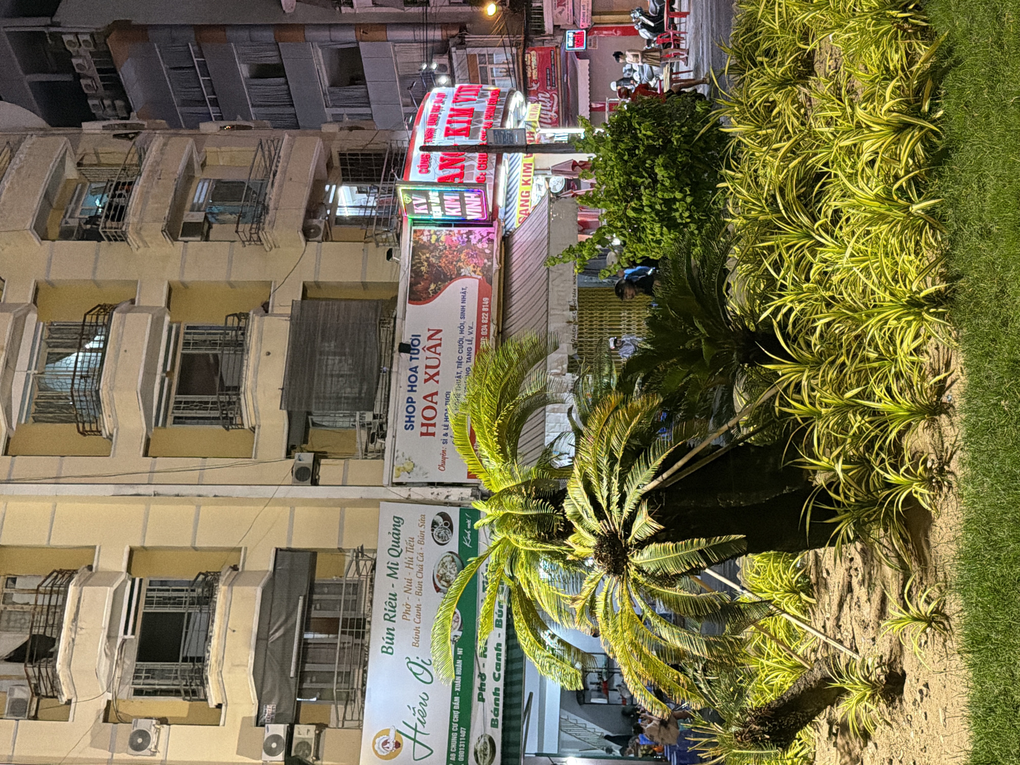 This media file captures a nighttime street scene in Nha Trang Ward, Vietnam. The focus is on a row of buildings with glowing neon signs, suggesting a commercial area. The signs advertise various businesses, including "SHOP HOA TƯƠI HOA XUÂN" (Fresh Flower Shop Xuan), "Hiếu Oi" (a restaurant specializing in Bún Riêu, Mì Quảng, Phở, and other noodle dishes), and "SANG KIM" which appears to be related to "VINH CUNG AN". The signs are predominantly in Vietnamese.
In the foreground, lush green plants and a palm tree are illuminated, creating a contrast with the urban backdrop. The overall scene suggests a lively evening in a Vietnamese city, likely a hub for dining and shopping. People are visible in the background, some sitting at tables outside restaurants and others on motorbikes, indicating ongoing activity. The lighting from the signs and streetlights casts a warm glow, typical of an urban setting at night. There are also air conditioning units visible on the building facades, hinting at the tropical climate. The combination of commercial signage, plant life, and human presence paints a picture of everyday life in a Vietnamese urban environment after dark.