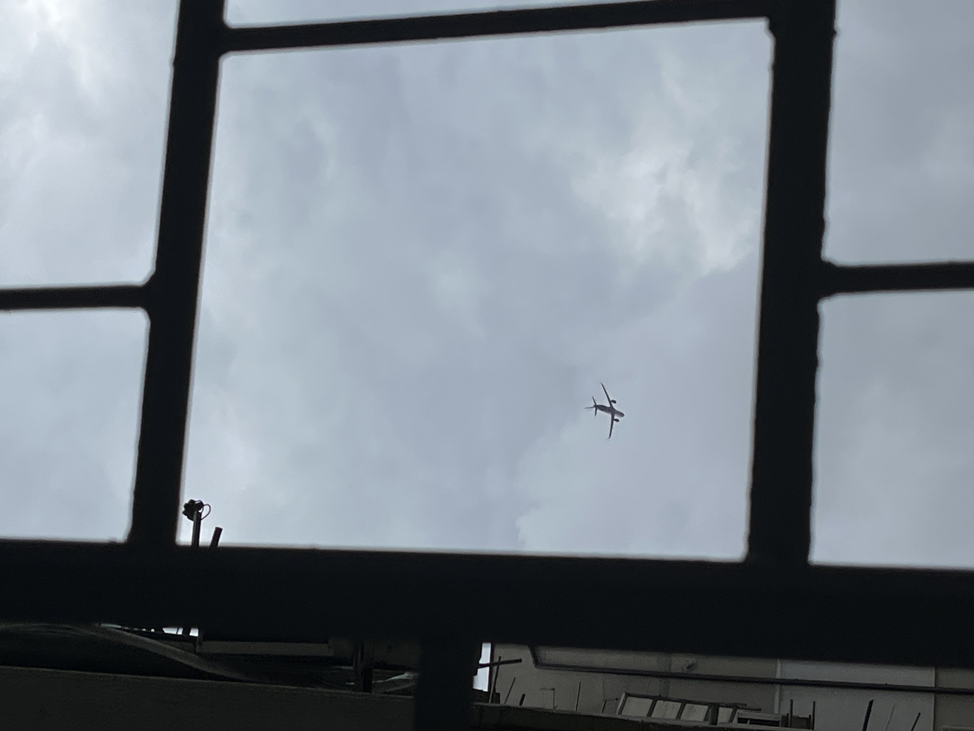 An airplane is silhouetted against a cloudy sky, seen through a window with black bars. The airplane is small and distant, with its wings spread. The sky is overcast with gray clouds. In the background, to the left, a portion of a building is visible, also silhouetted. The image appears to have been taken from indoors, looking out. The scene is devoid of people. There are no contextual clues to indicate the location is Varna, Bulgaria.