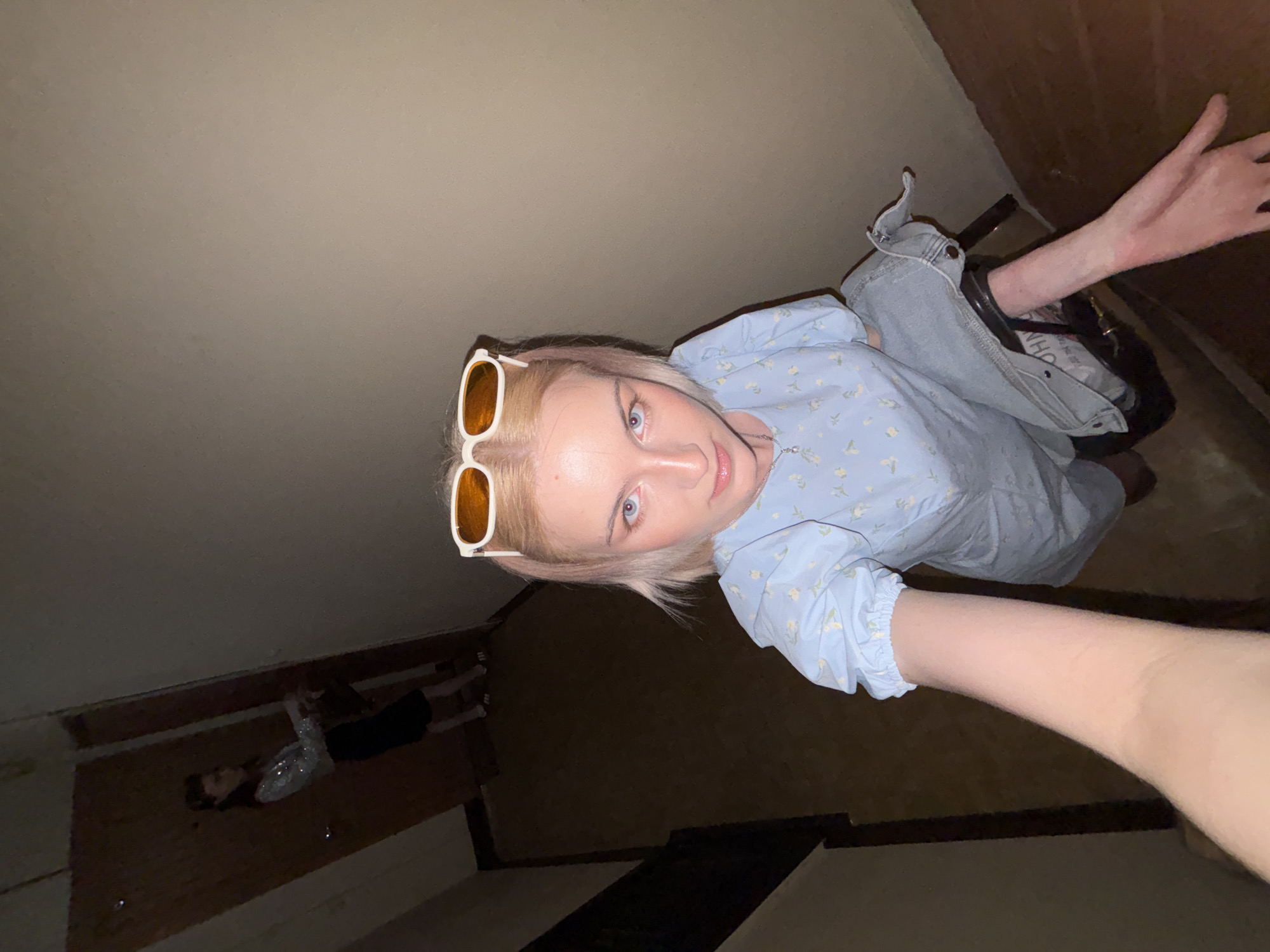 An indoor scene in Vsevolozhsk, Russia, depicts two females within a stairwell or hallway environment. In the foreground, a light-skinned female with blonde hair and light blue eyes looks directly at the camera. She wears a light blue short-sleeved dress with small yellow floral patterns and a thin silver necklace. White-framed sunglasses with amber lenses are pushed onto her head. Her left arm extends towards the camera, holding a light blue denim garment and a dark handbag. In the background, a second female with dark hair, wearing a shimmering silver long-sleeved top and a dark skirt, stands near a dark wooden door on a lower landing or hallway. The walls are a plain light brown, and the floor is a darker brown. A dark banister or handrail is partially visible on the left side of the frame. The lighting is moderate, illuminating the foreground subject more brightly than the background.