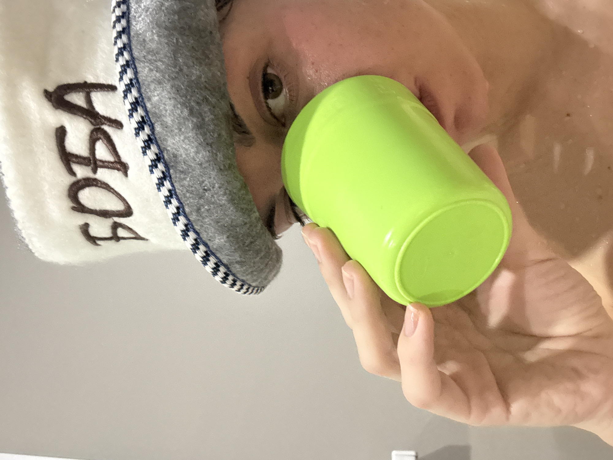 An indoor, close-up shot features a person wearing a white and grey woolen hat, possibly a sauna hat, with visible Cyrillic text embroidered on it. The text appears to be "ЧФОБА". The person's face is partially visible, showing one eye looking towards the viewer, and the nose and mouth area. Their skin appears wet and slightly flushed, suggesting they are in a warm or humid environment, such as a sauna or bath, or have recently left one. The person is holding a bright neon green plastic cup to their mouth, in the act of drinking. A hand with neatly trimmed nails is visible holding the cup. The background is indistinct but appears to be a light-colored wall or tiled surface. The overall activity suggests rehydration or relaxation in a bath or sauna setting, in Ust-Ilimsk, Russia.
