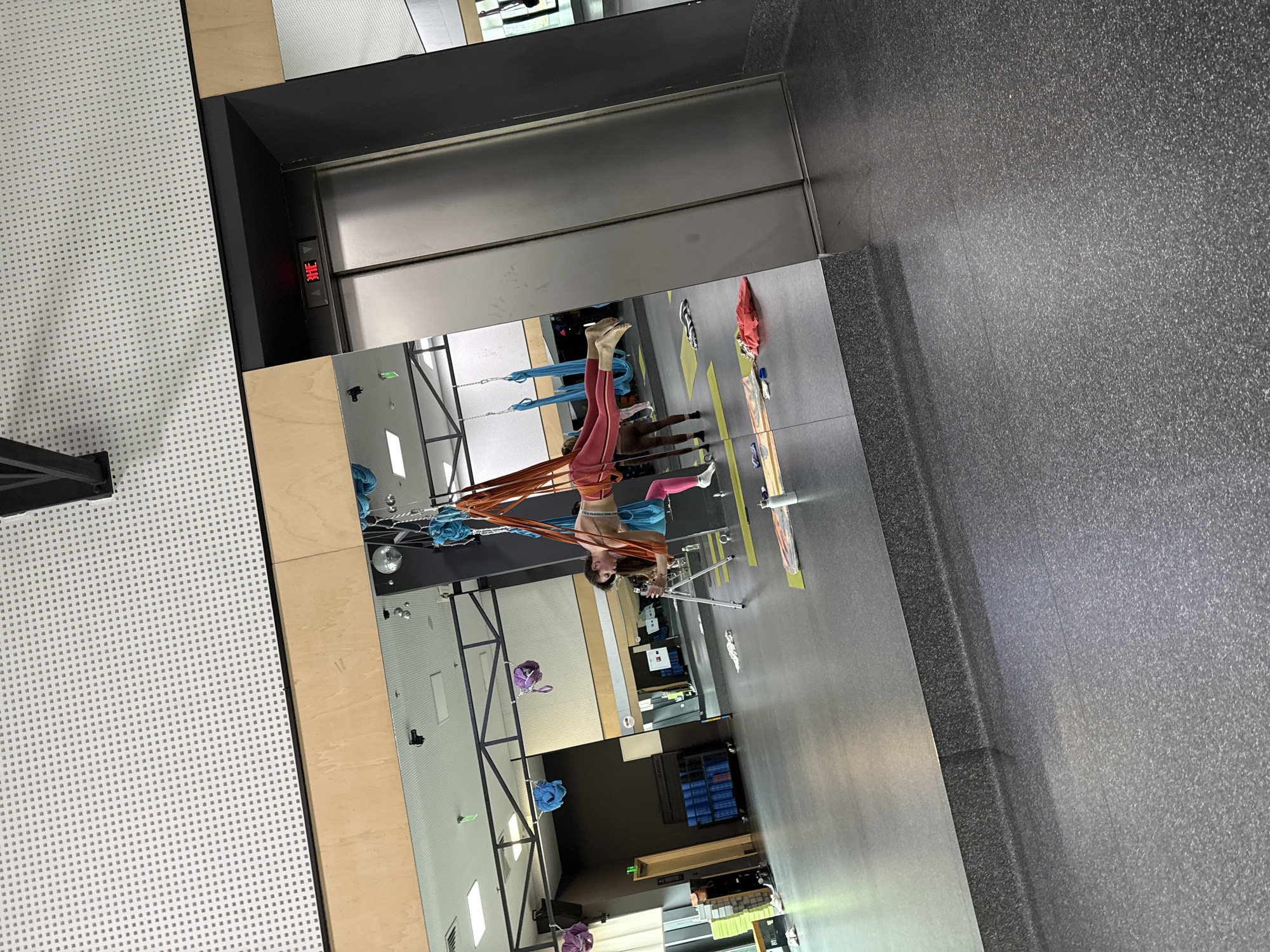 Here is a forensic description of the image:
The image depicts a reflection in an elevator mirror, showing a woman suspended upside down from aerial silks in what appears to be a fitness studio. She is wearing athletic clothing, with the silks wrapped around her torso and legs. Various yoga mats and fitness equipment are visible in the background of the studio. The elevator itself is a standard model with a digital floor display. The surrounding area includes walls with a textured, grid-like pattern and a gray, speckled floor. The city and specific location are unknown.