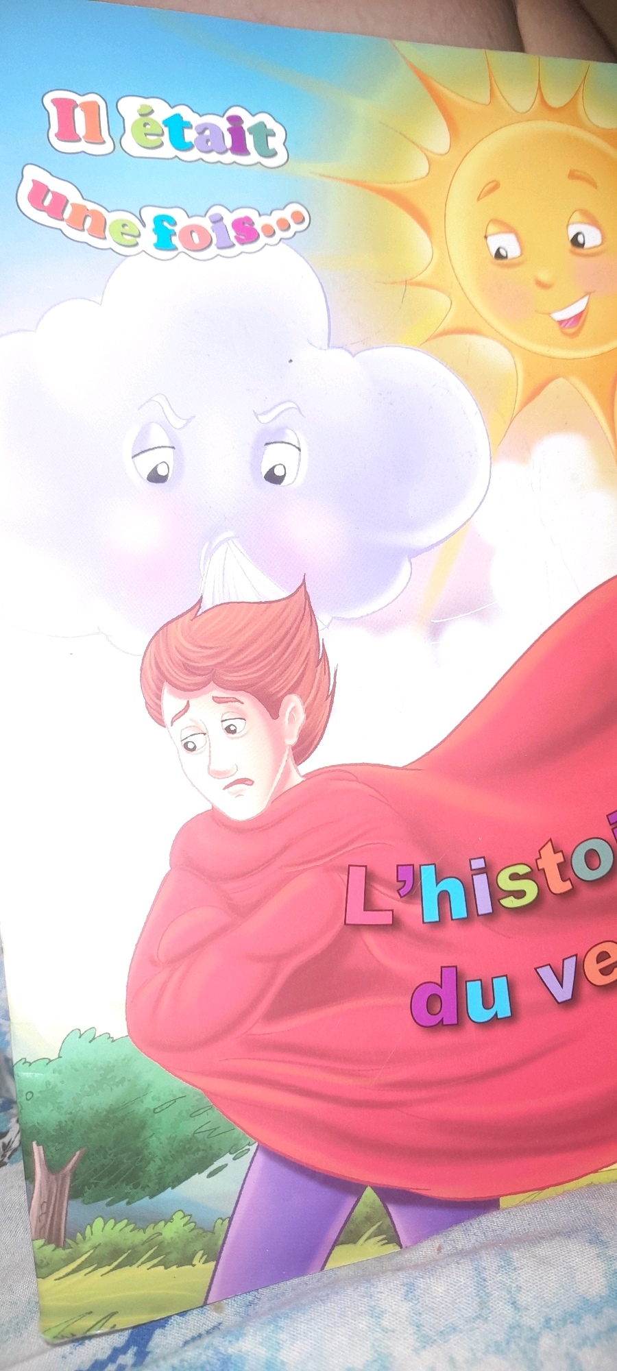 The image is a close-up, angled shot of a children's book cover. The cover features a cartoon illustration with a bright blue sky and sunshine on the right. In the center, a stylized white cloud with an angry expression hovers above a young boy with red hair and a distressed look on his face. The boy is wearing a large, red cape. Text in colorful, bold letters reads "Il était une fois..." in the top left corner. Below the boy, also in colorful letters, are the words "L'histori du ve". The bottom of the image shows a simplified landscape with green grass and trees. The book cover appears to be lying on a blue and white patterned fabric. The image does not contain any information or visual cues that would allow for identification of a location in Marrakesh, Morocco.