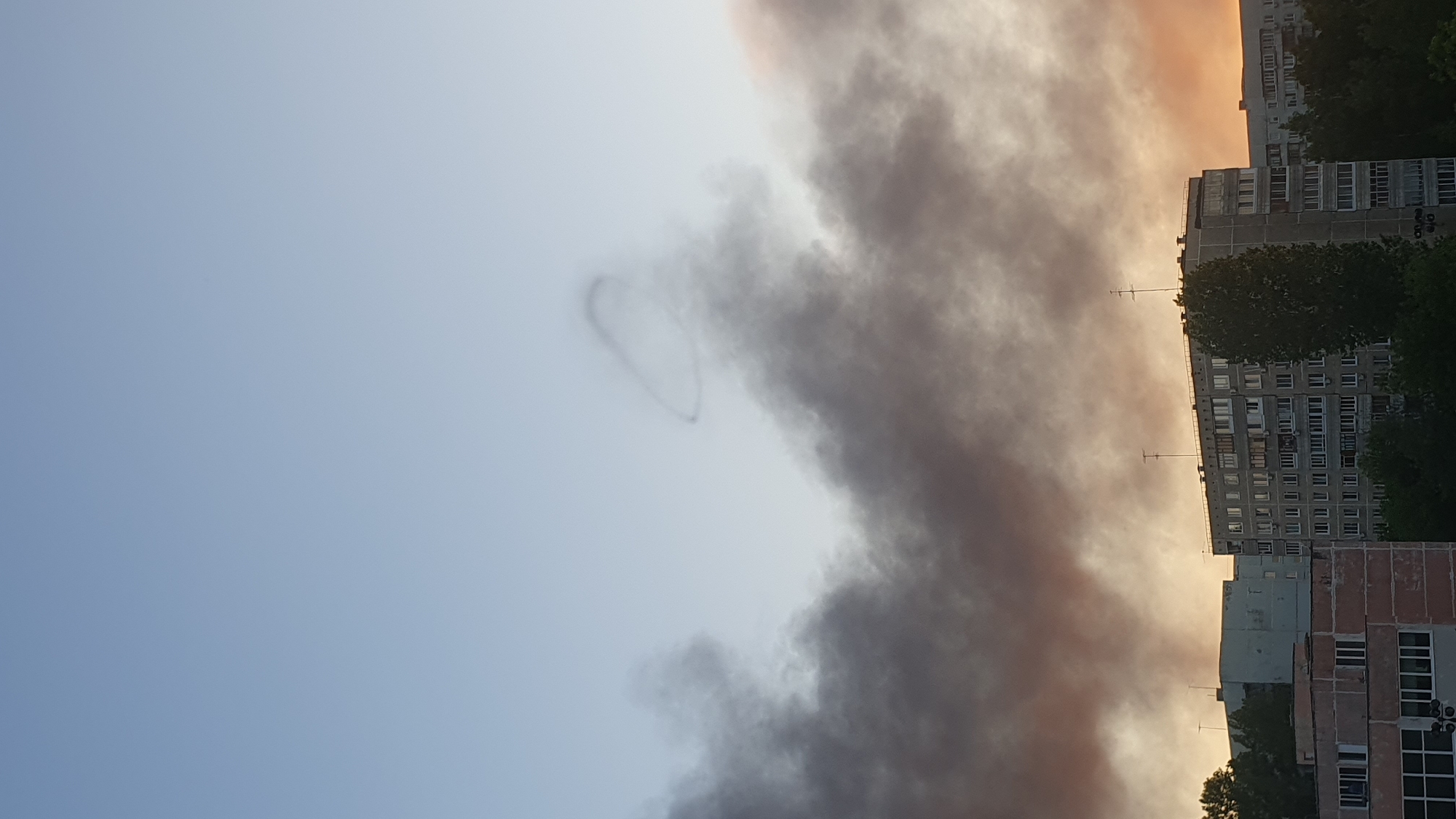 The image captures a sky over Ulyanovsk, Russia. The sky is partially obscured by a large, dark cloud formation, possibly smoke. To the right, residential buildings are visible, suggesting an urban environment. In the clear area of the sky, the contrail of an airplane is visible, looped in the form of a heart.