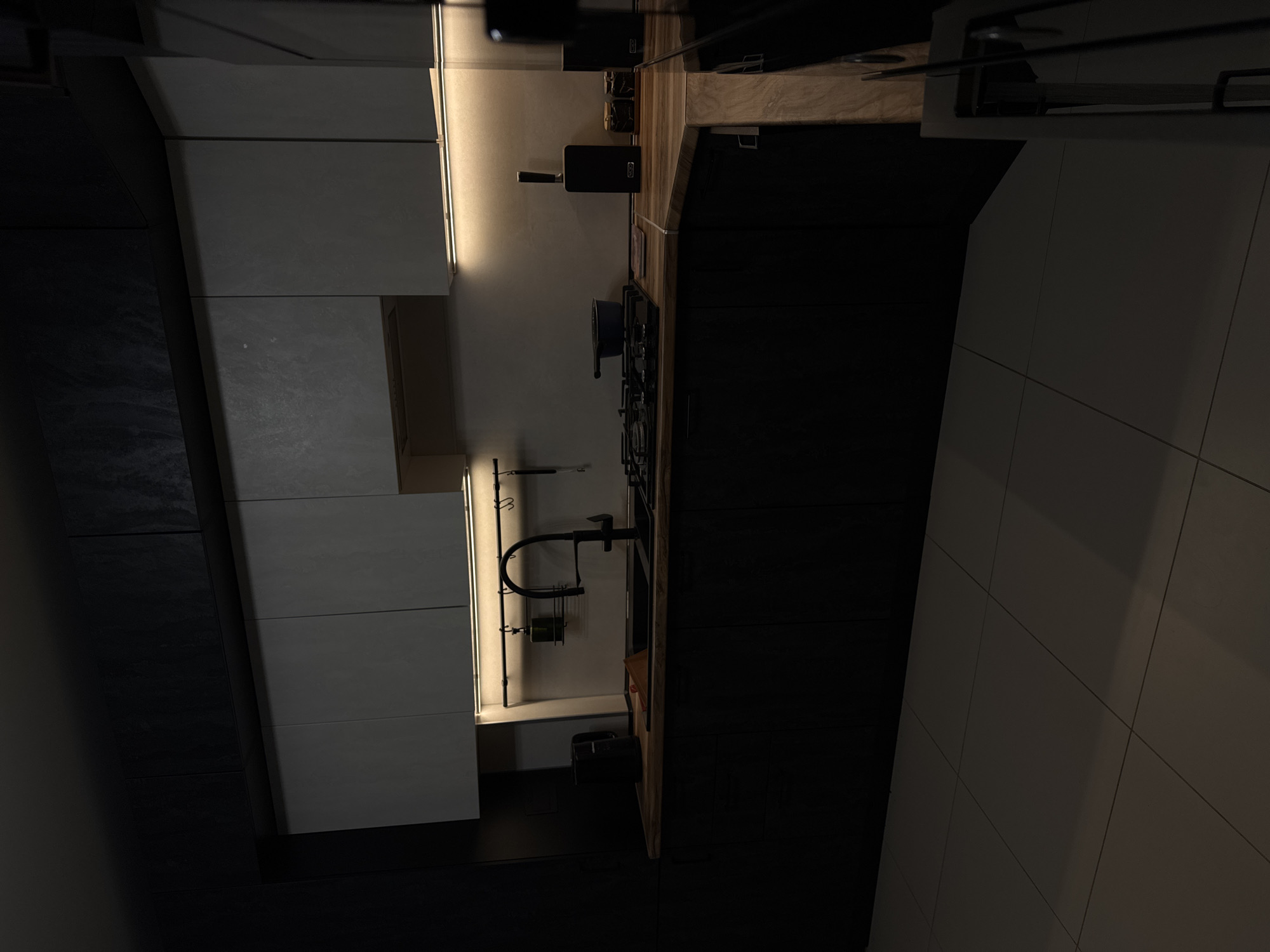 The image depicts a modern, dark-themed kitchen, likely located in Mikhaylovsk, Russia. The scene is dimly lit, with warm LED strip lighting under the upper cabinets casting a glow on the countertop. The kitchen features dark, matte lower cabinets and a contrasting lighter-colored upper cabinet structure. A black faucet with a high arc is prominent over a black sink. To the right of the sink, on the wooden countertop, is a wooden cutting board and a blue pot on a black stovetop. Further to the right, a knife block with a single knife is visible, along with two small, dark containers, possibly for spices or condiments. The floor is tiled with large, light-colored square tiles. The overall impression is one of a clean, minimalist, and contemporary kitchen space.