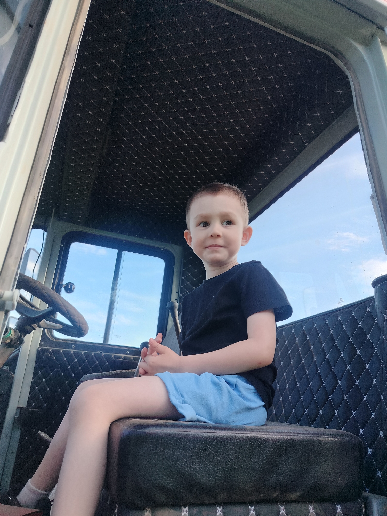 A young child sits in the driver's seat of a vehicle with a black quilted interior. The child wears a dark t-shirt and light blue shorts. A steering wheel is visible to the left, and windows show a sky with light clouds. The vehicle appears to be a tractor. The location is unknown.
